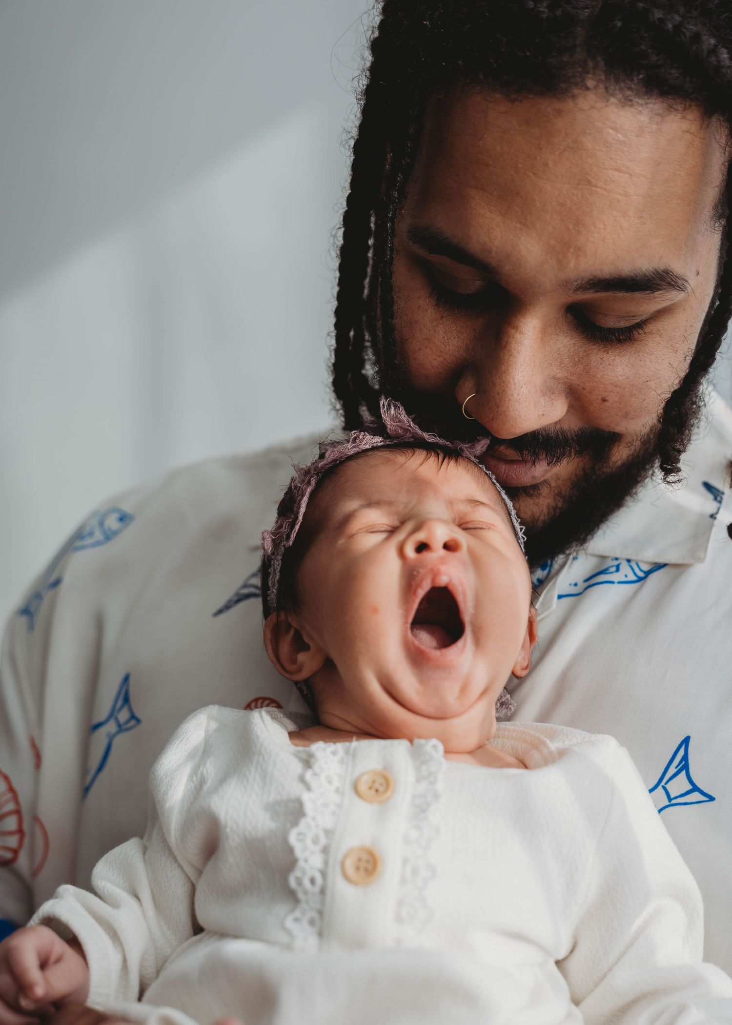 A father with dreadlocks and nose ring holds his yawning newborn baby dressed in white with a floral headband.