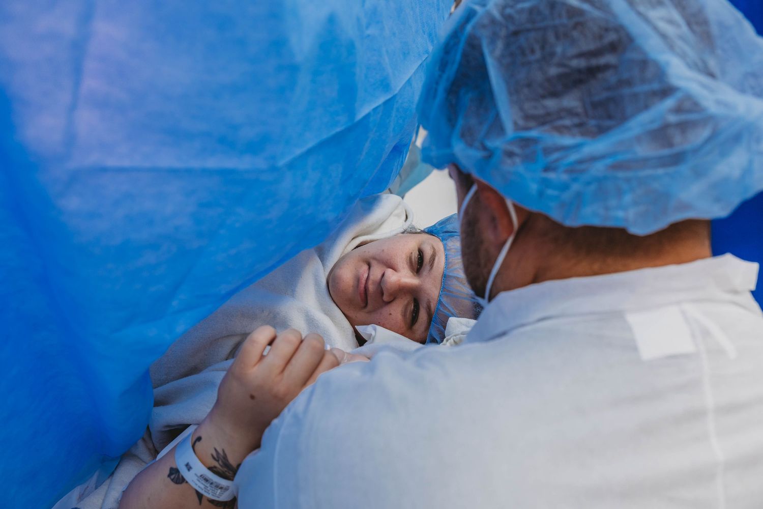 Smiling new mother lies under blue surgical drapes holding her newborn as a masked partner in scrubs leans in during a C-section delivery.