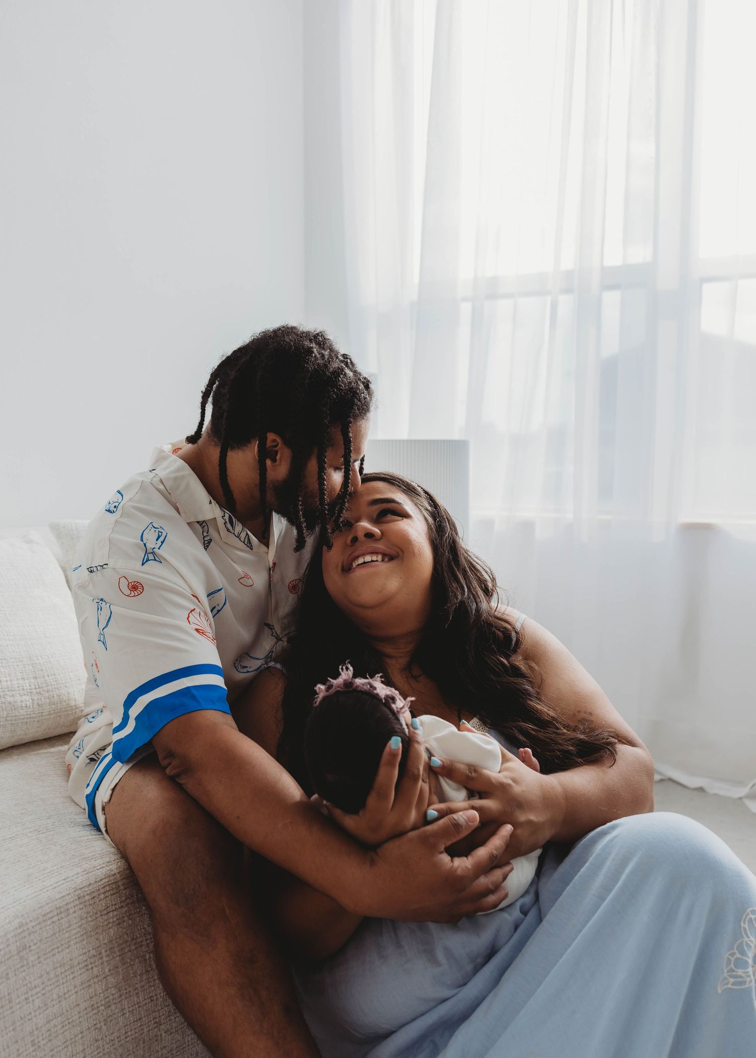 A couple sits on a couch cradling their newborn baby, sharing a tender moment in a bright, airy studio during their newborn session.