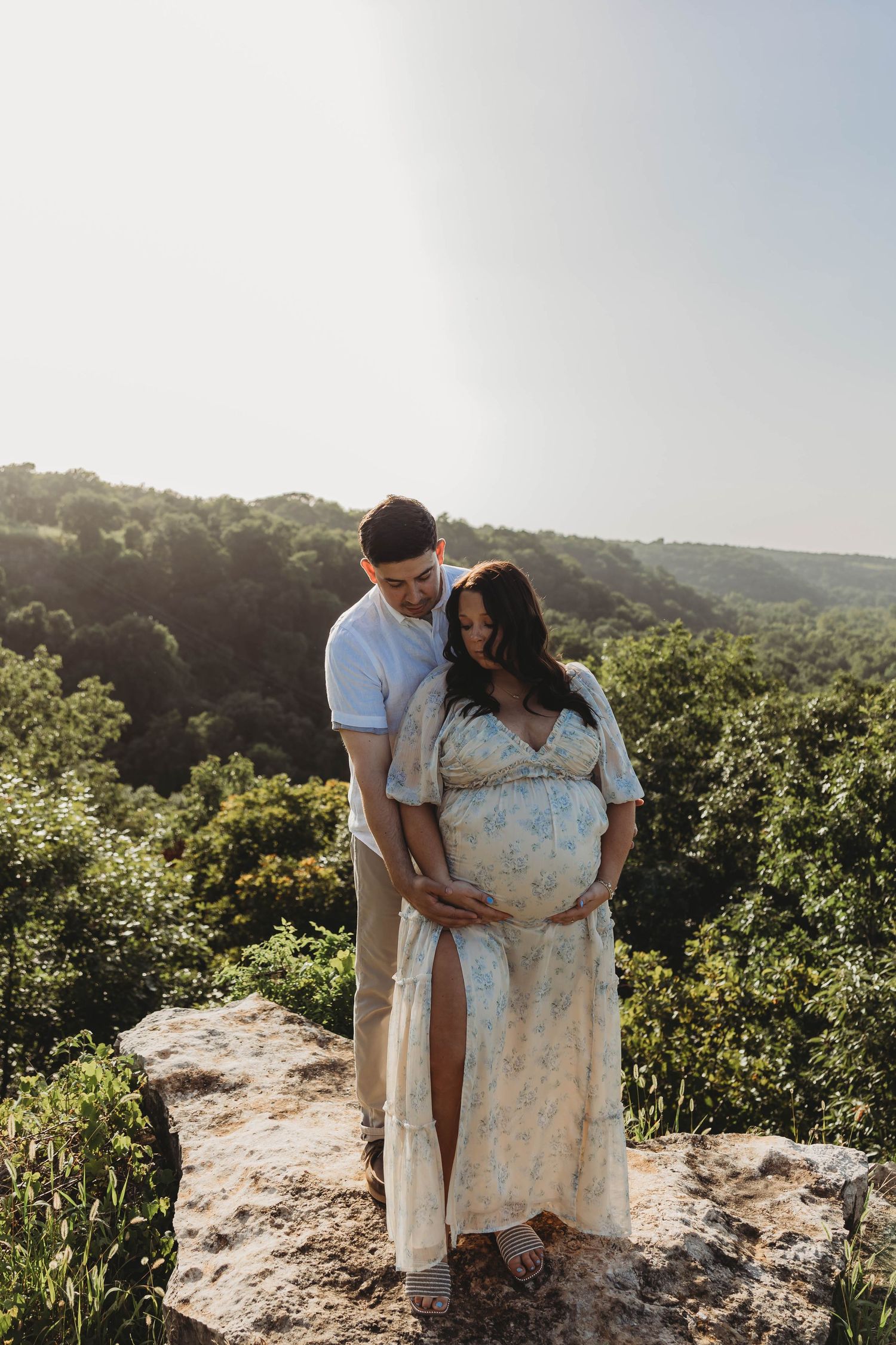 Pregnant couple embracing on rocky overlook with lush forest backdrop.