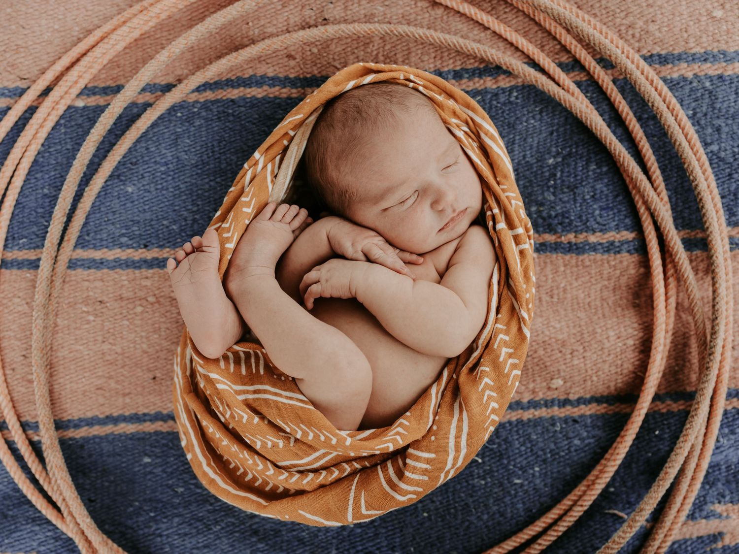 Sleeping newborn baby curled in a western blanket on a striped blue horse blanket with a rope border in Big Cabin Oklahoma with Keely King