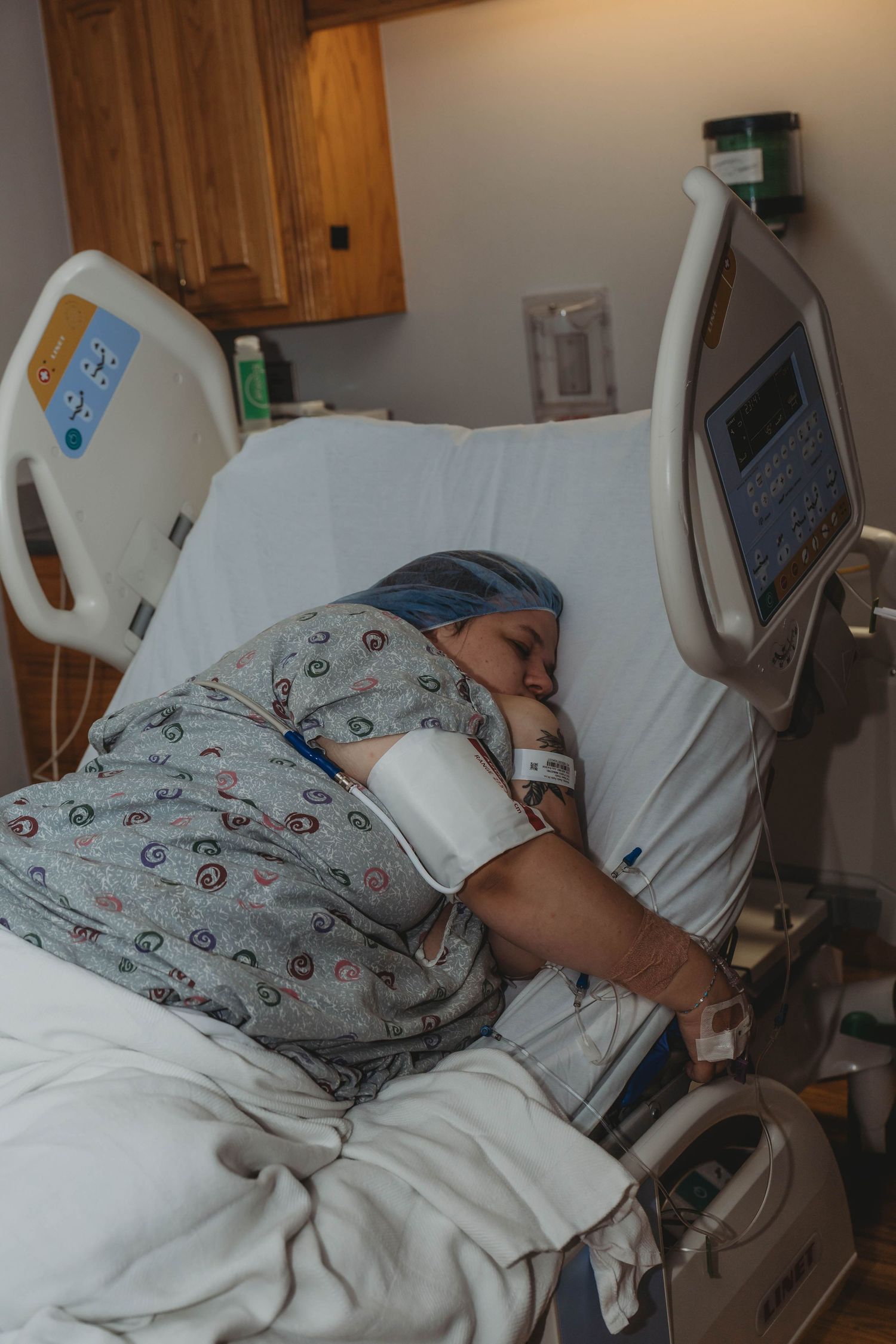 A patient with blue hair rests in a hospital bed wearing a gown, with a blood pressure cuff and IV attached.