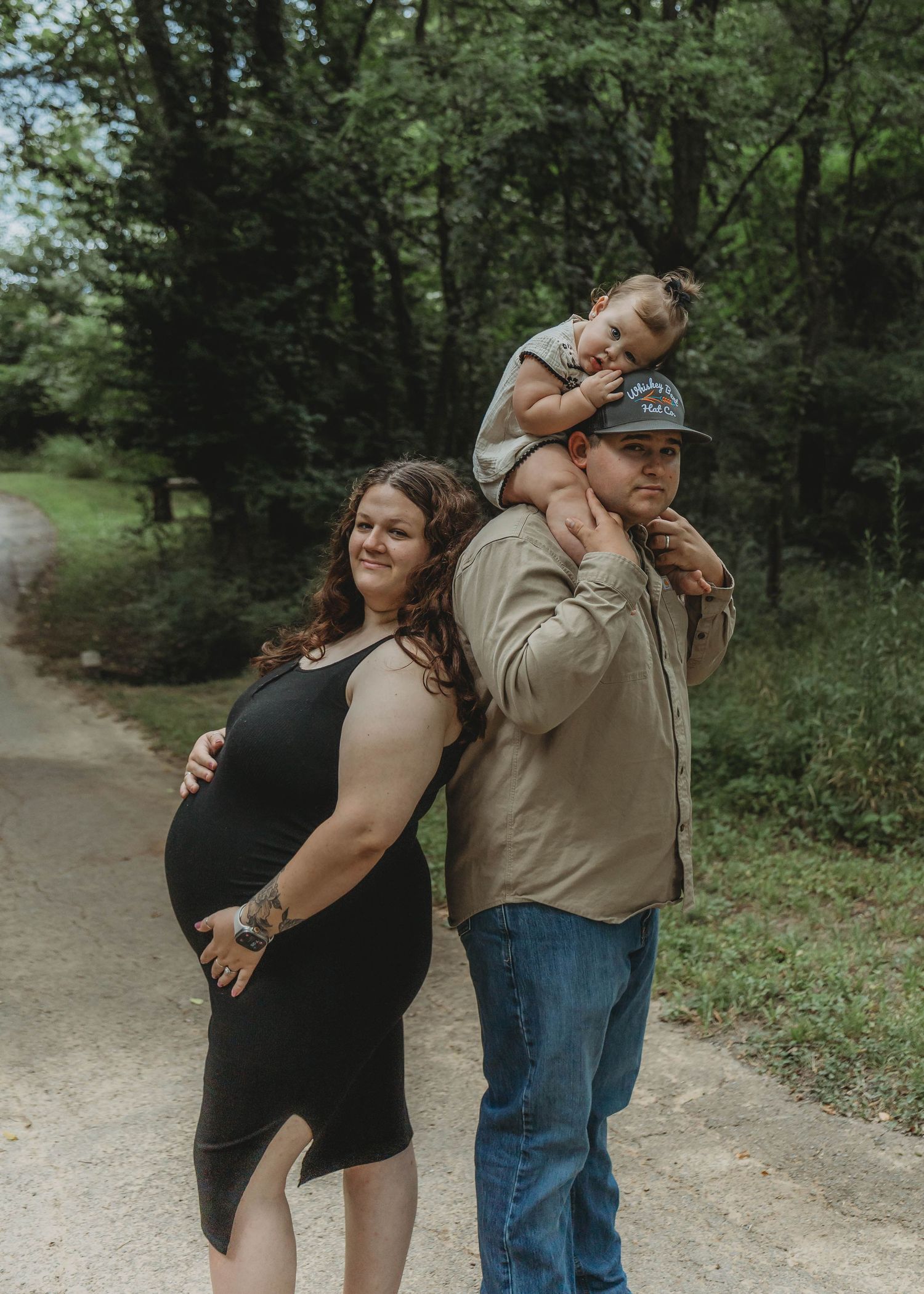 Pregnant woman in black dress poses with partner carrying toddler on shoulders on a wooded path.