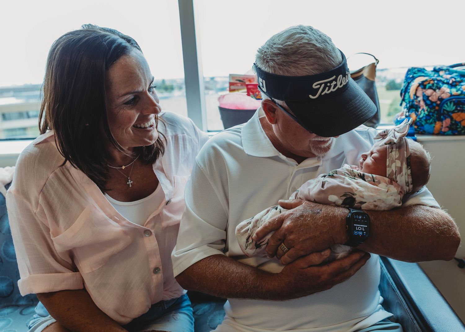 Proud grandparents smiling joyfully while holding their newborn grandchild in hospital room.