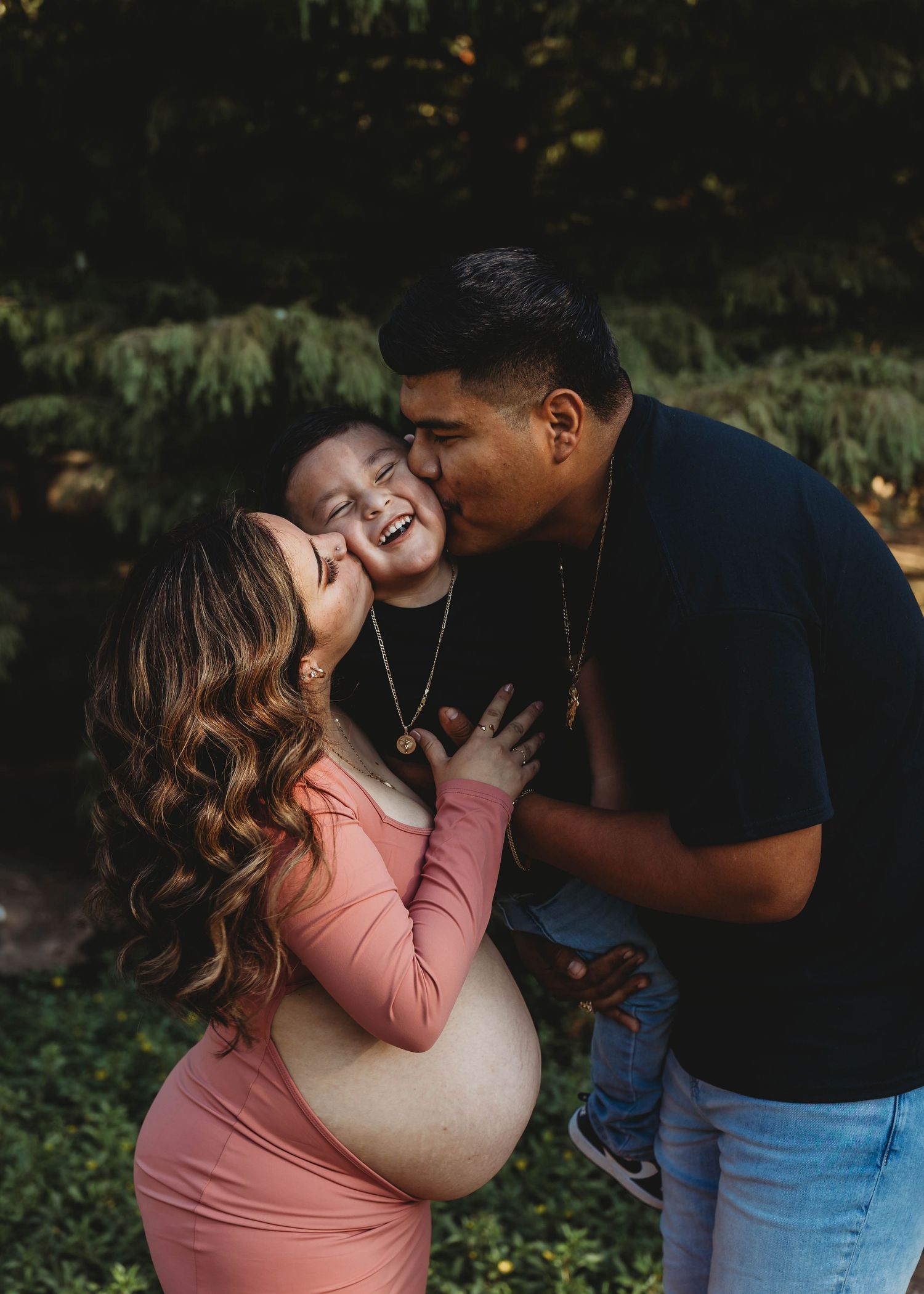 Pregnant woman in pink dress smiles as man kisses child between them outdoors in lush green setting.