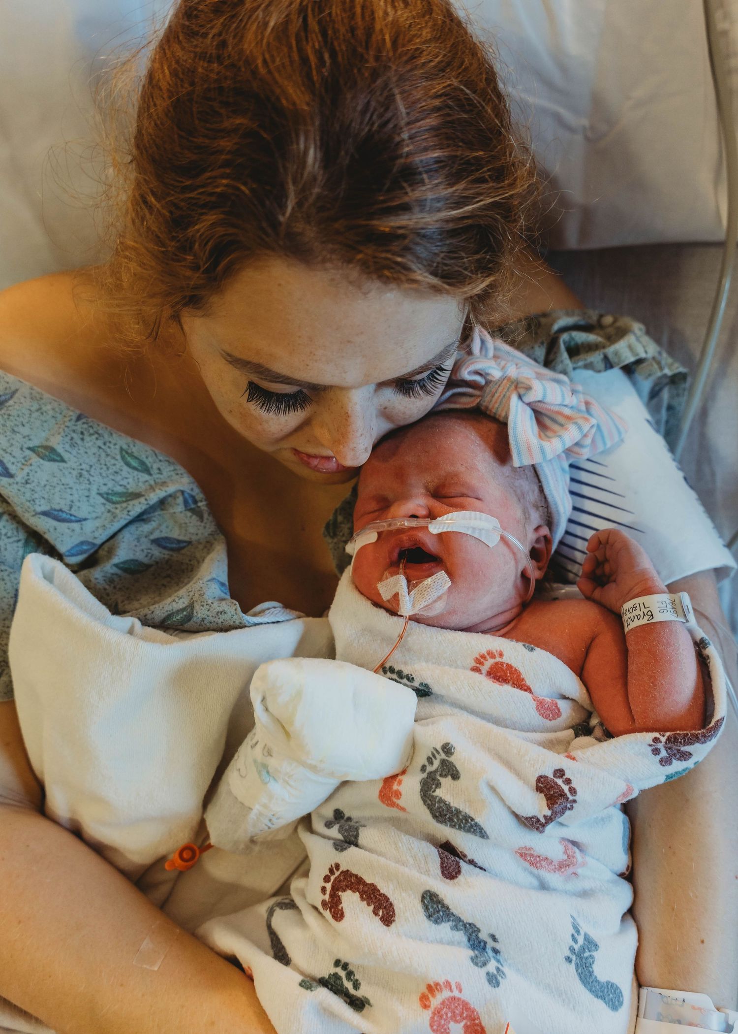 New mother in hospital gown tenderly holds and gazes at her newborn baby wrapped in a footprint blanket.