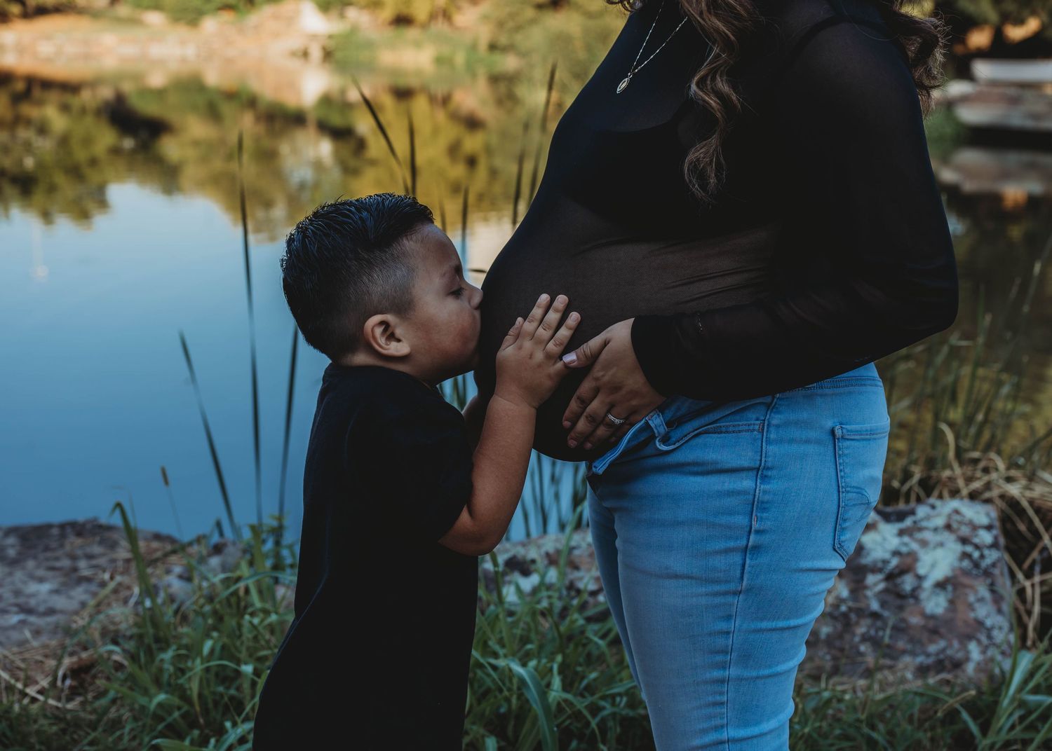 A young boy kisses his pregnant mother's belly near a serene lake during a maternity photo session outdoors.