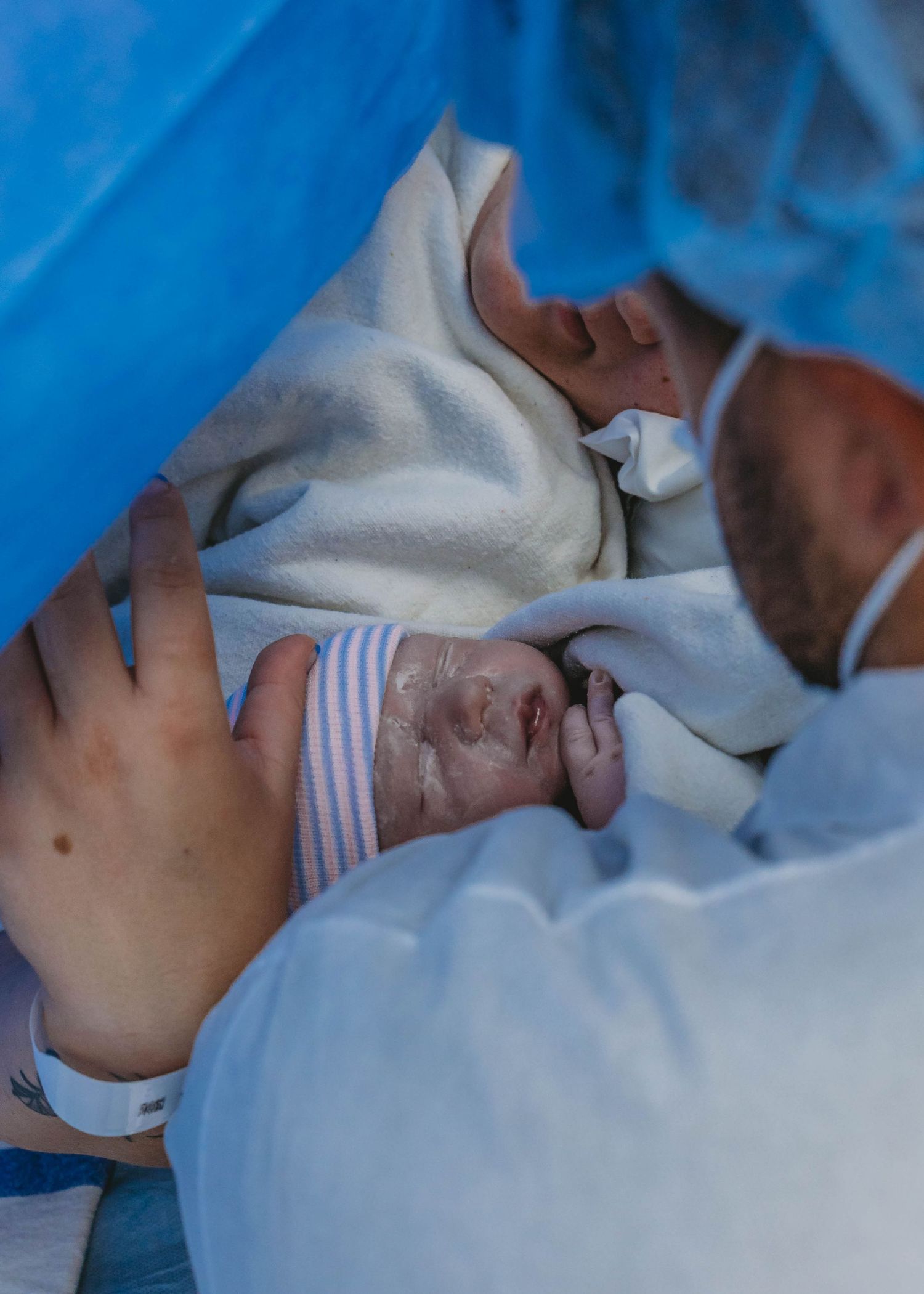 Newborn baby wrapped in blanket held by parent wearing surgical mask in hospital delivery room.