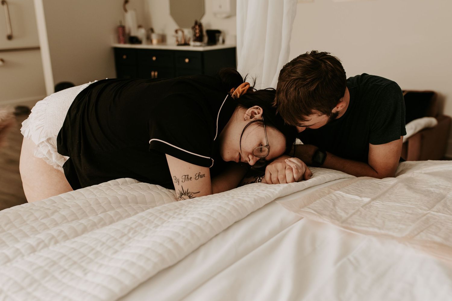 Couple in matching black shirts sharing an intimate moment lying together on a white bed in a cozy bedroom.