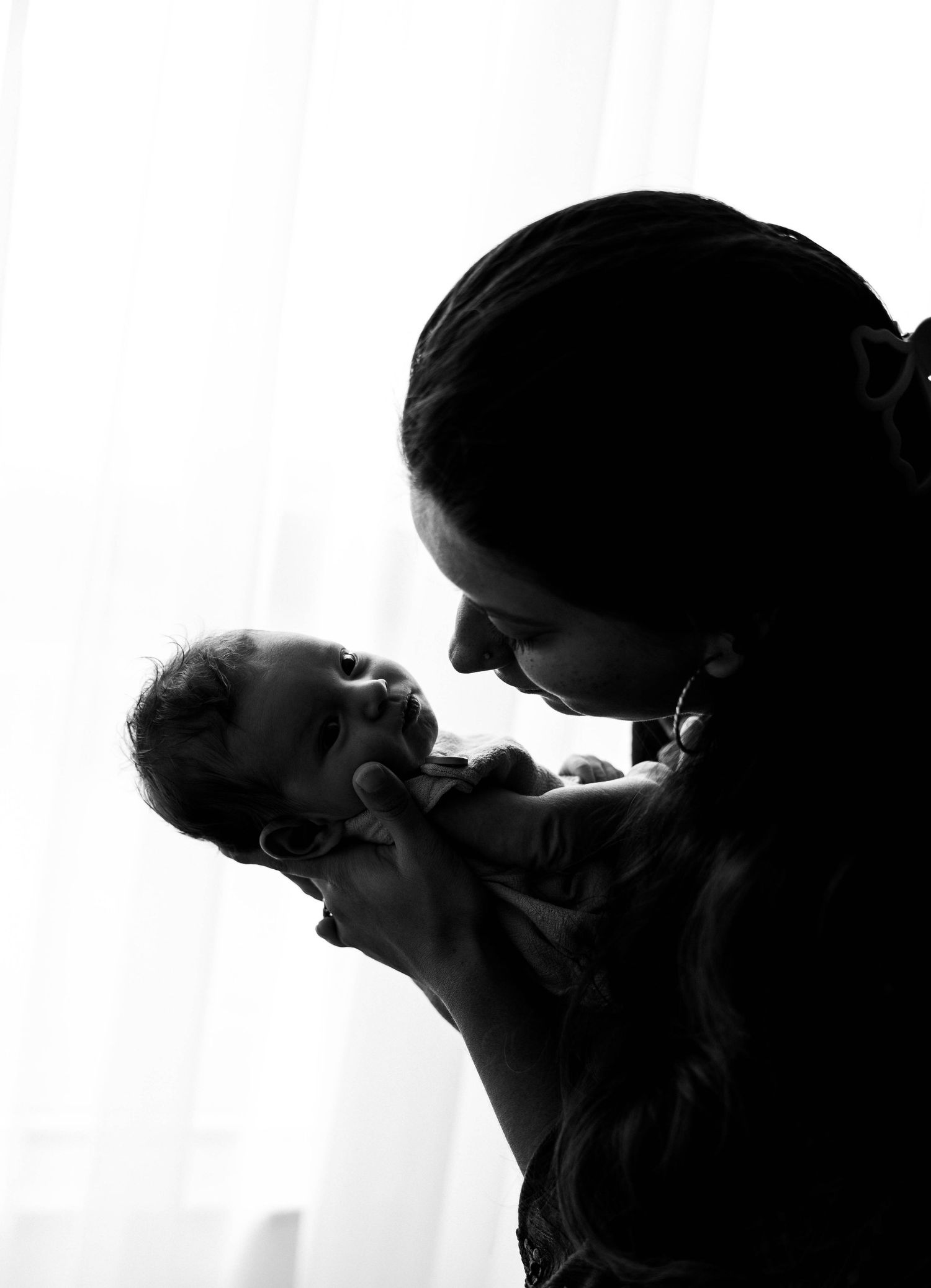 Black and white silhouette of a mother tenderly holding her newborn baby near a bright window.
