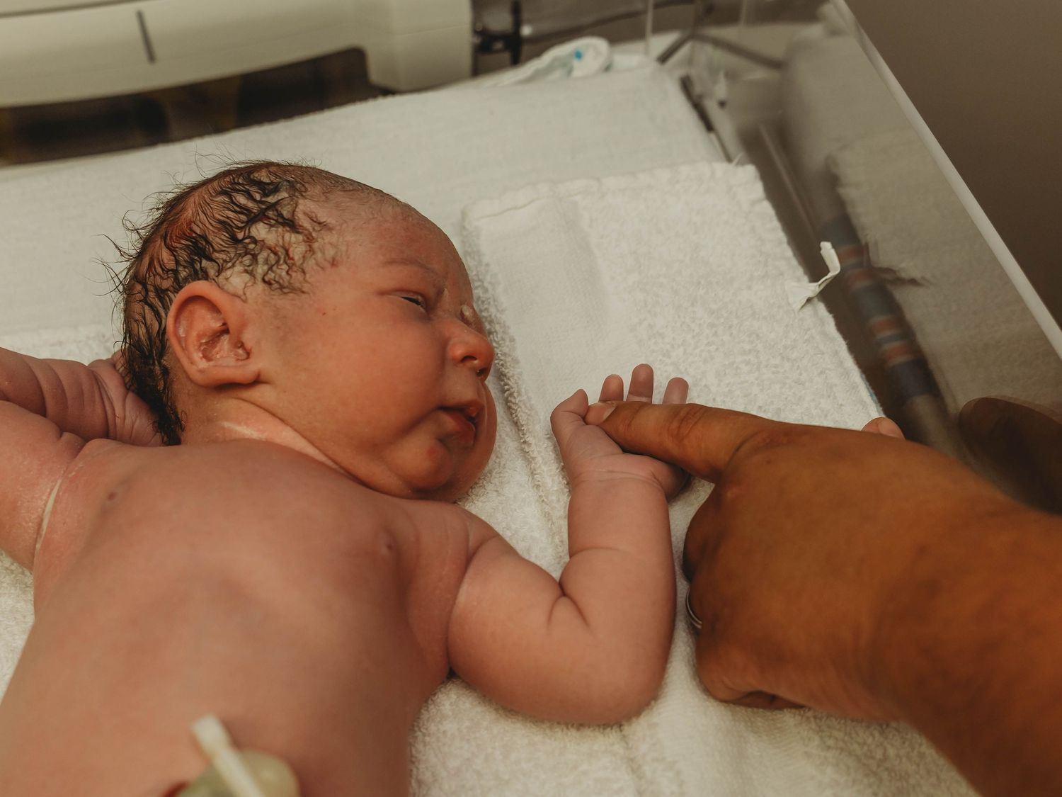 Newborn baby with damp hair lying on a white towel in a hospital bed while an adult hand gently holds the infant's tiny fingers.