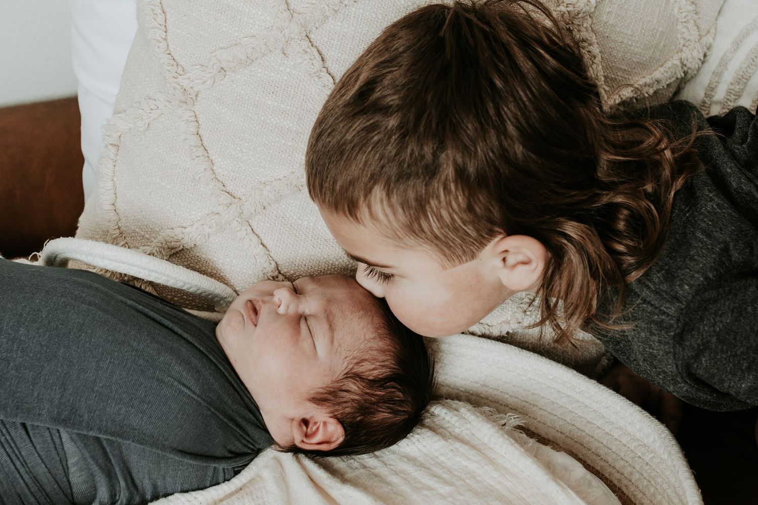 Older child tenderly kissing a sleeping newborn baby lying on a soft white blanket at home.
