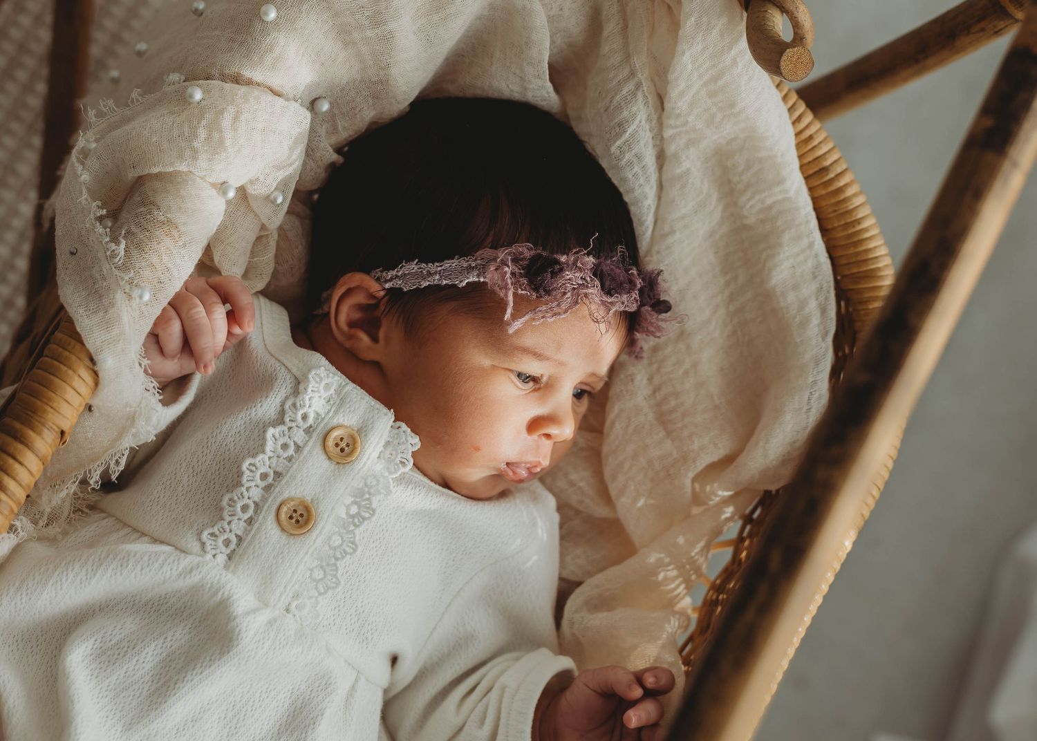 Newborn baby peering peacefully in a wicker bassinet, wearing a white outfit and delicate floral headband in a studio by Keely King Photo.
