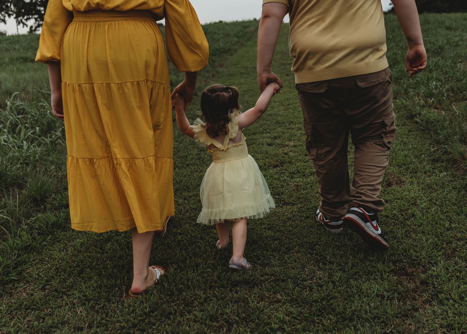 Pregnant mother and father holding toddler daughters hands while walking through green grass during maternity session.
