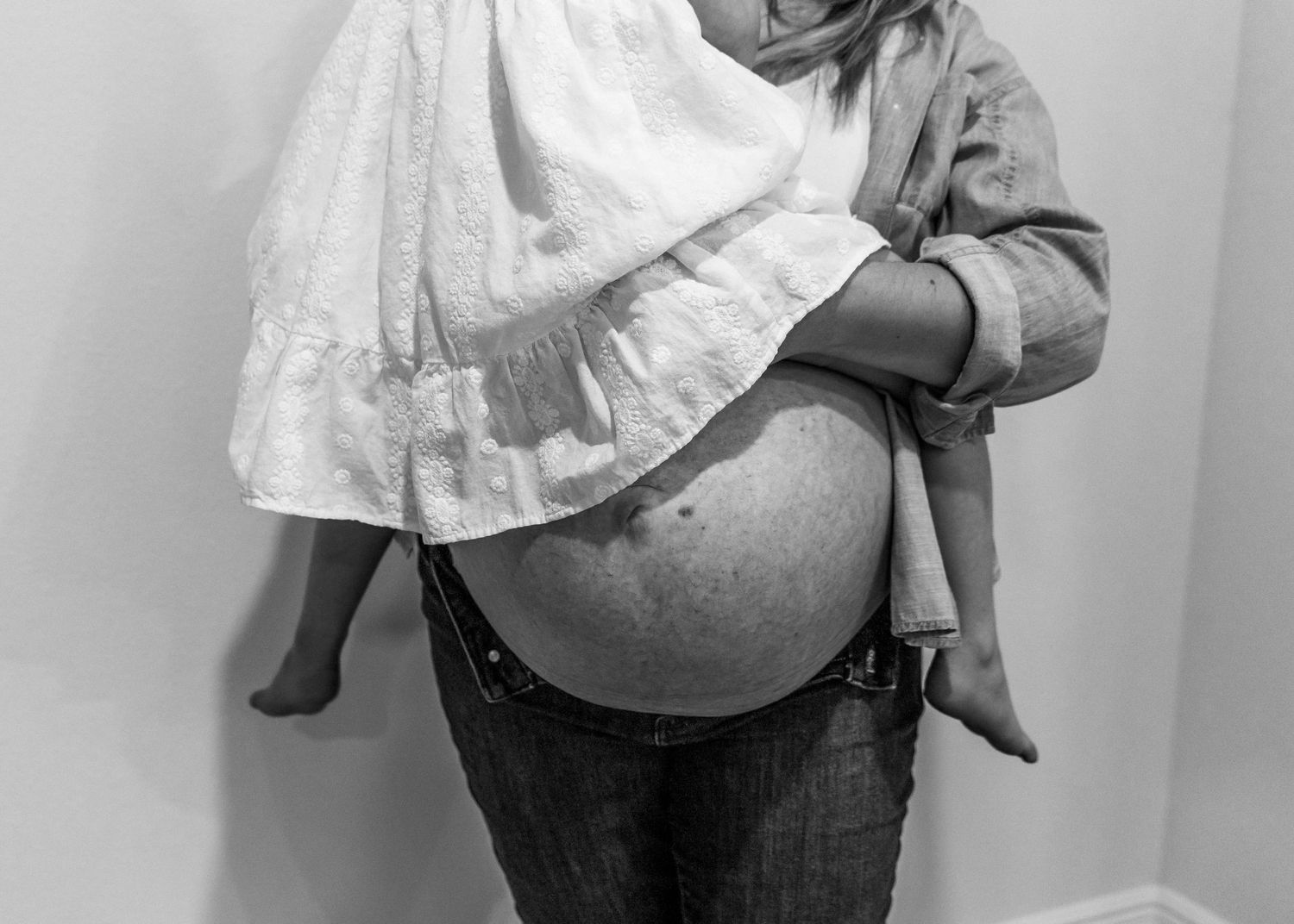 Black and white photo of a pregnant woman holding a child, showcasing her baby bump in jeans and an open shirt.