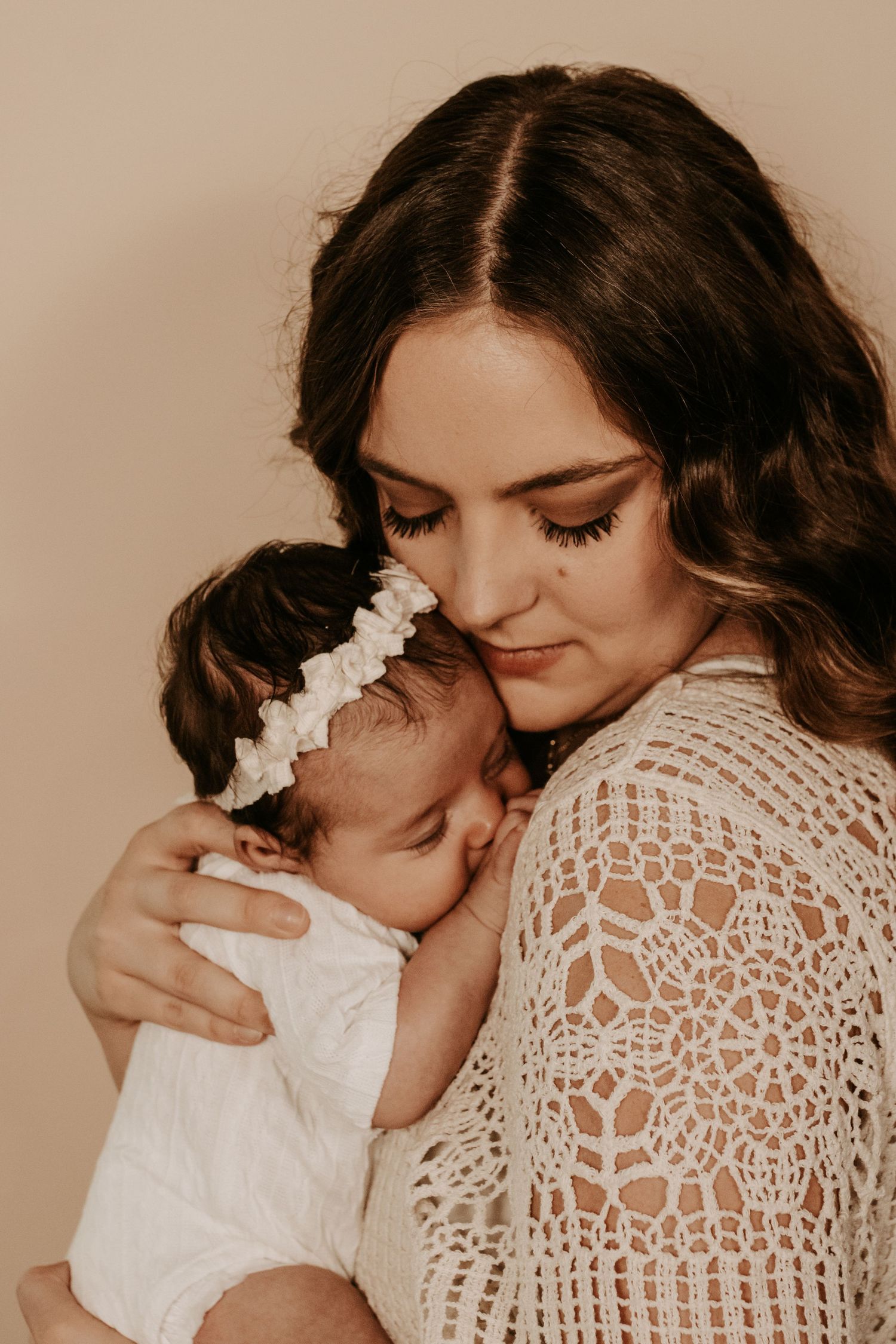 A young mother with dark hair tenderly holds her newborn baby girl in a white onesie and floral headband.