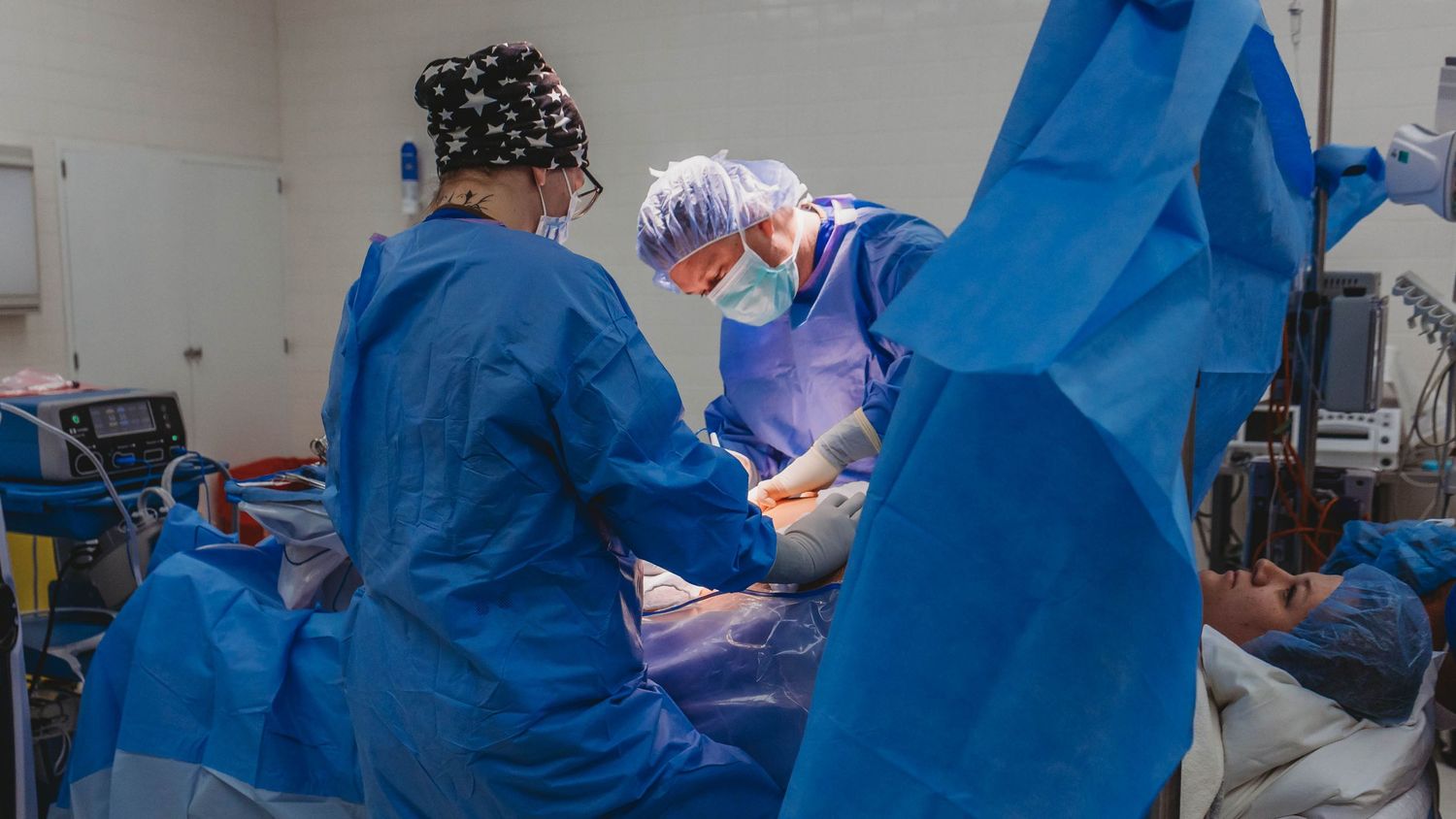 Two surgeons in blue gowns, masks and surgical caps perform a procedure on a patient in a bright, equipped operating room.