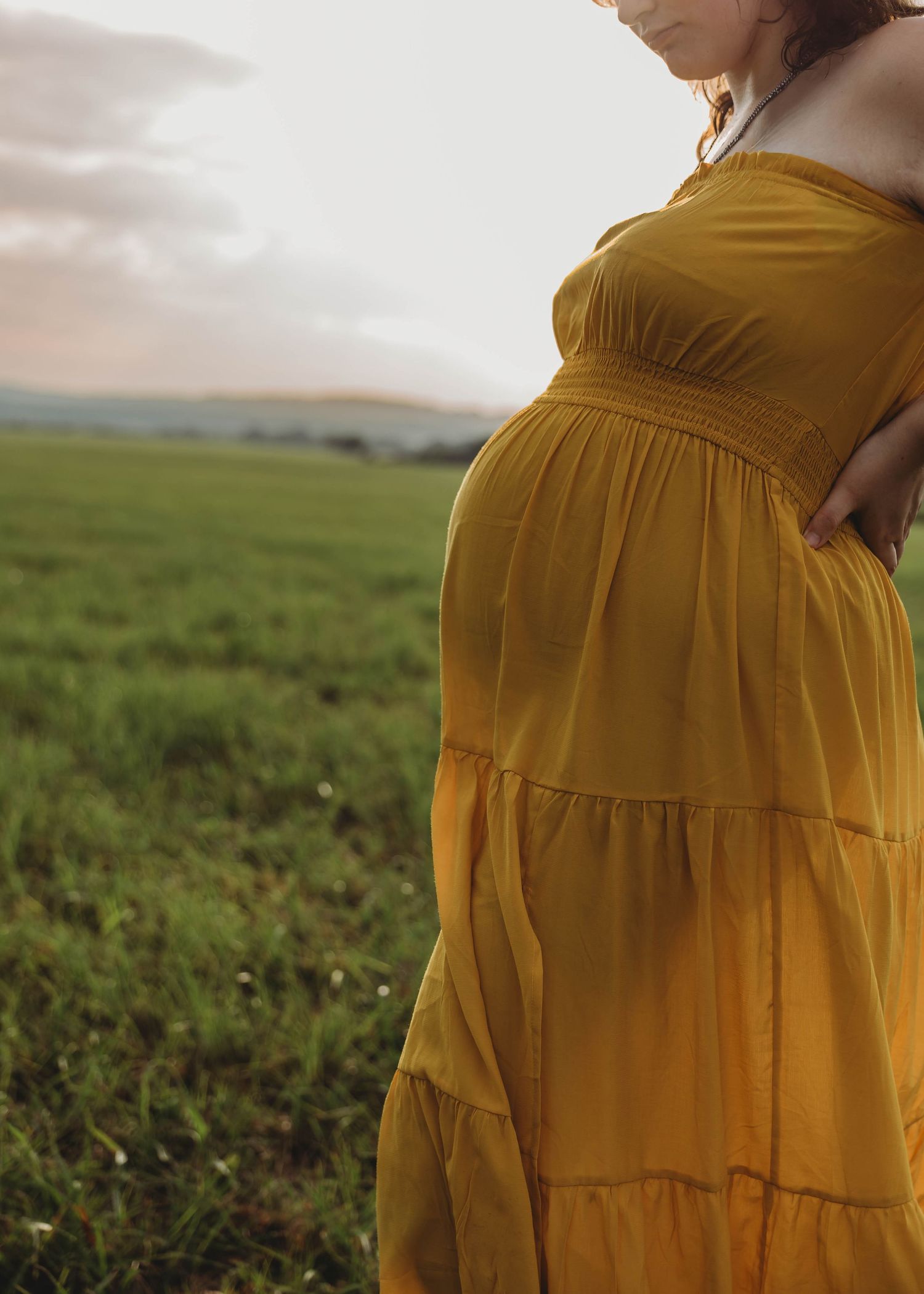 Pregnant woman in mustard yellow off-shoulder maxi dress standing in green field during golden hour maternity session.