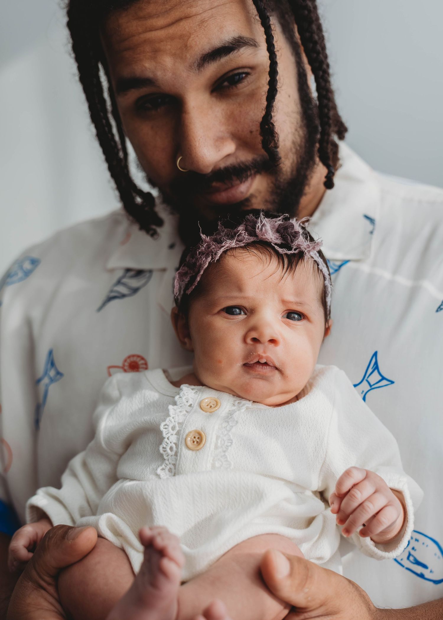 A father with dreadlocks and nose ring tenderly holds his newborn baby girl wearing a white outfit and floral headband.