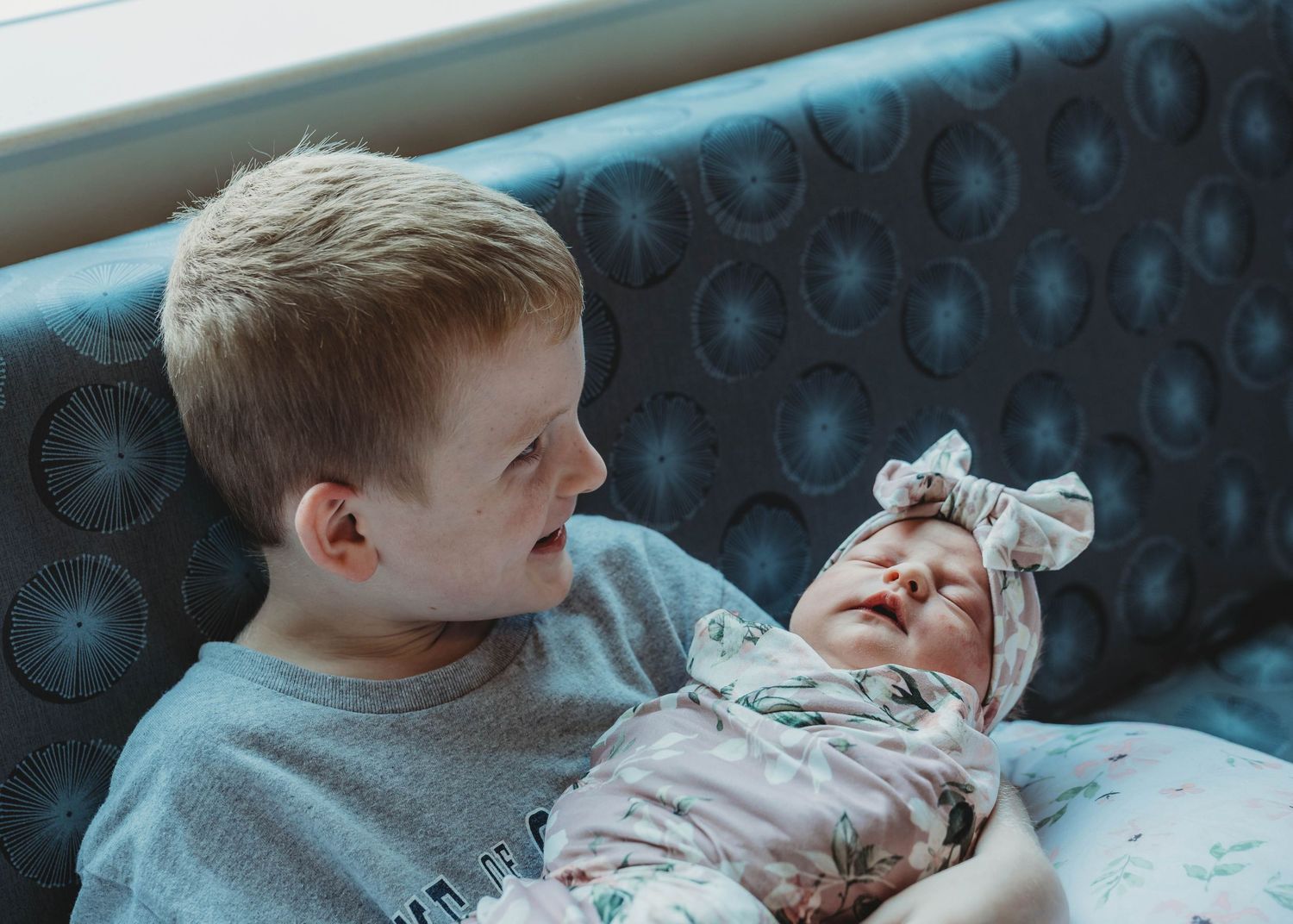 Young boy lovingly gazing at his newborn baby sister wrapped in a floral swaddle.