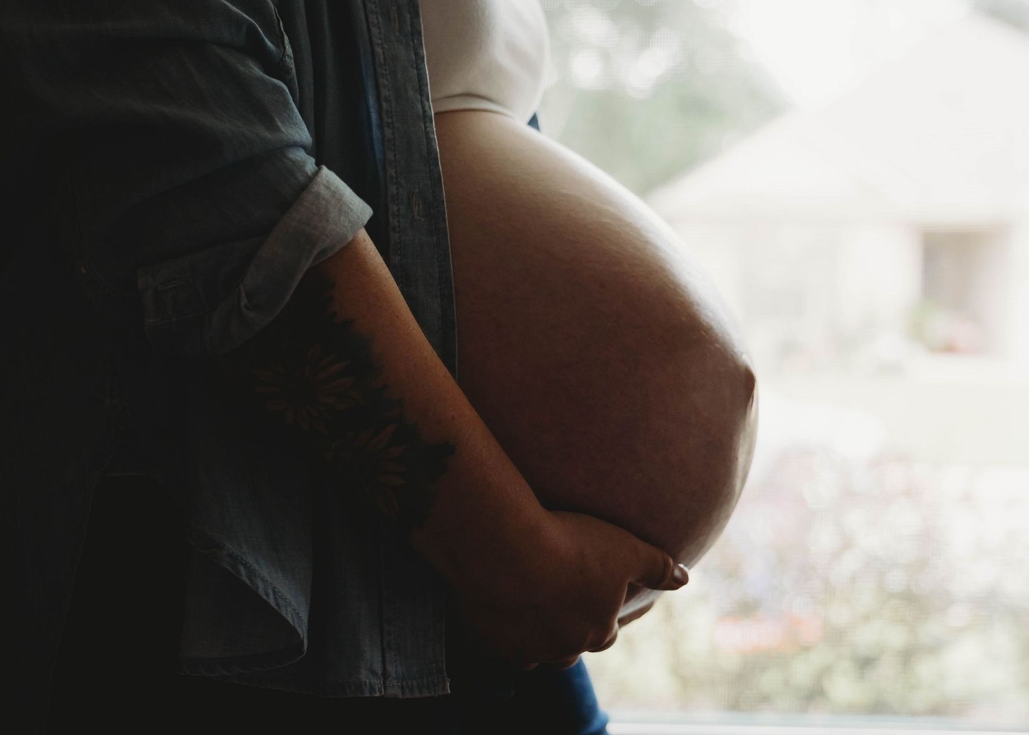 Pregnant woman cradling her baby bump, wearing a denim jacket, standing near a window with soft natural light.