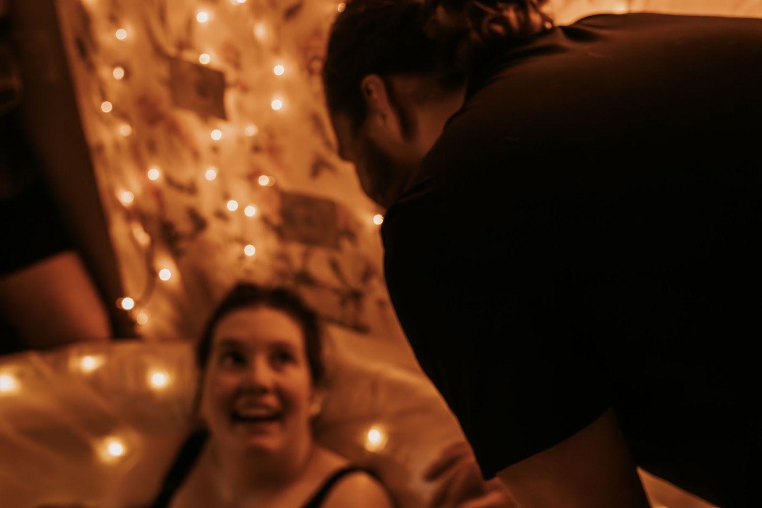 Two people share a joyful moment in a warmly lit room decorated with string lights in the background taken by Keely King Photography