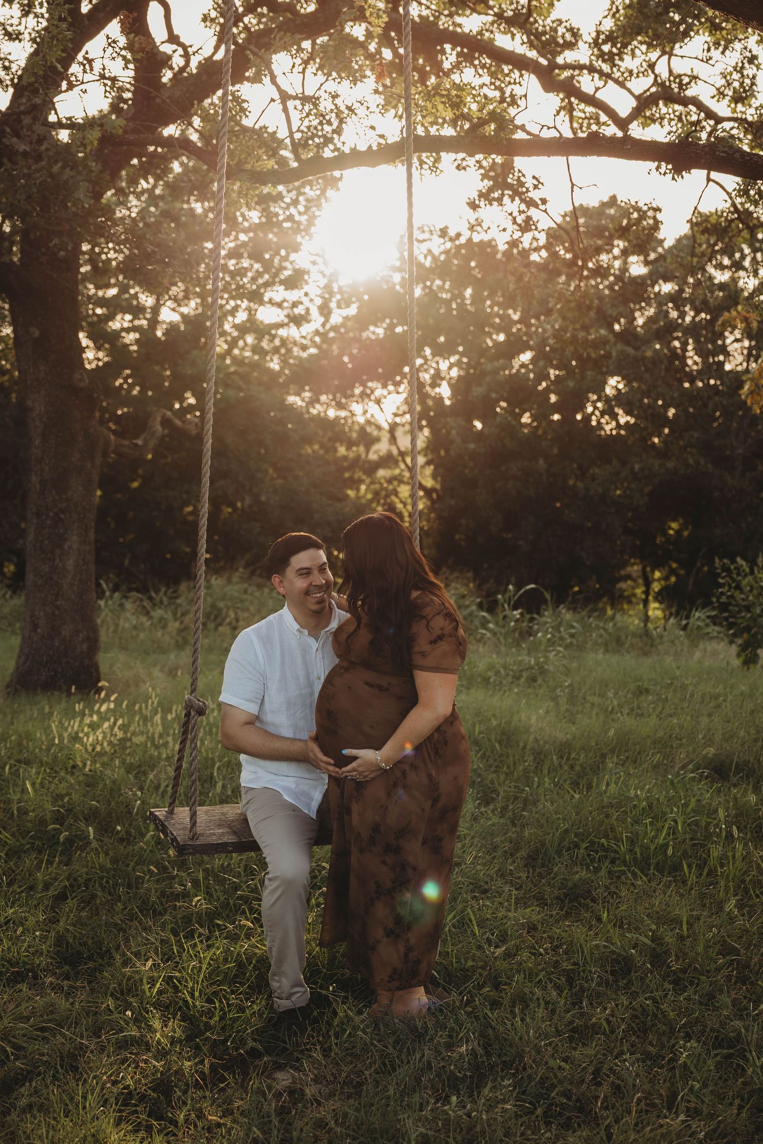 Couple sharing a tender moment by a rope swing in a sunlit field during maternity shoot.