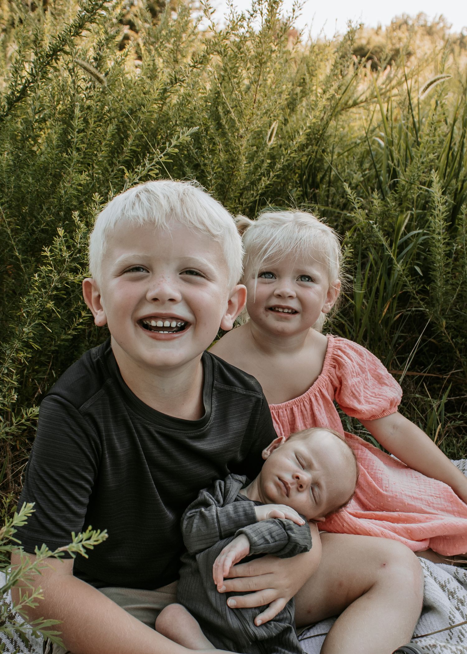 Two blonde siblings smile outdoors holding a sleeping newborn baby, surrounded by tall green grass in a warm sunset setting in Claremore, OK