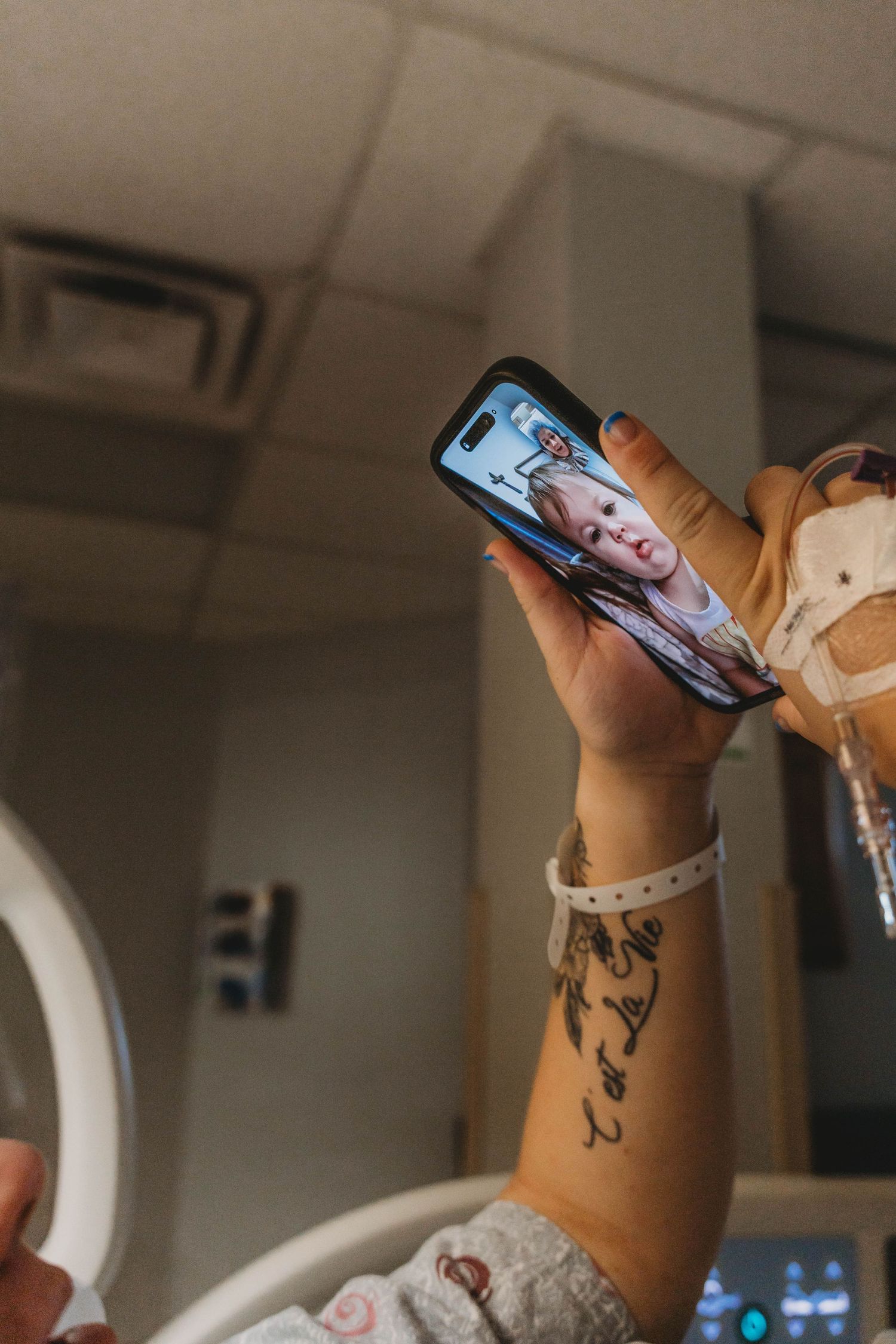 Hospital patient with an IV and ID bracelet holds up a smartphone showing a video call with a smiling toddler from a hospital bed.