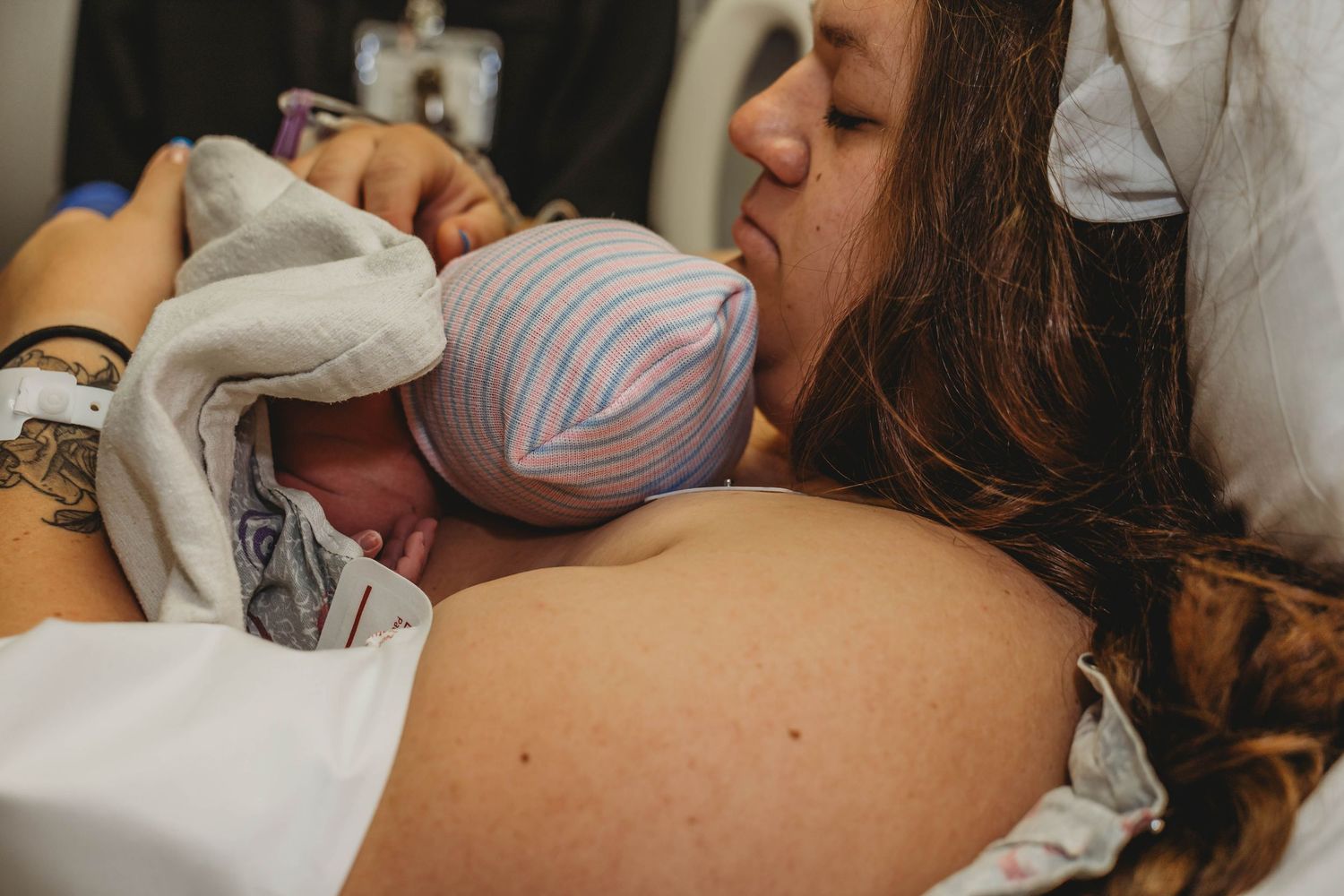 A new mother rests in a hospital bed holding her newborn, who wears a striped knit hat, skin-to-skin against her chest.