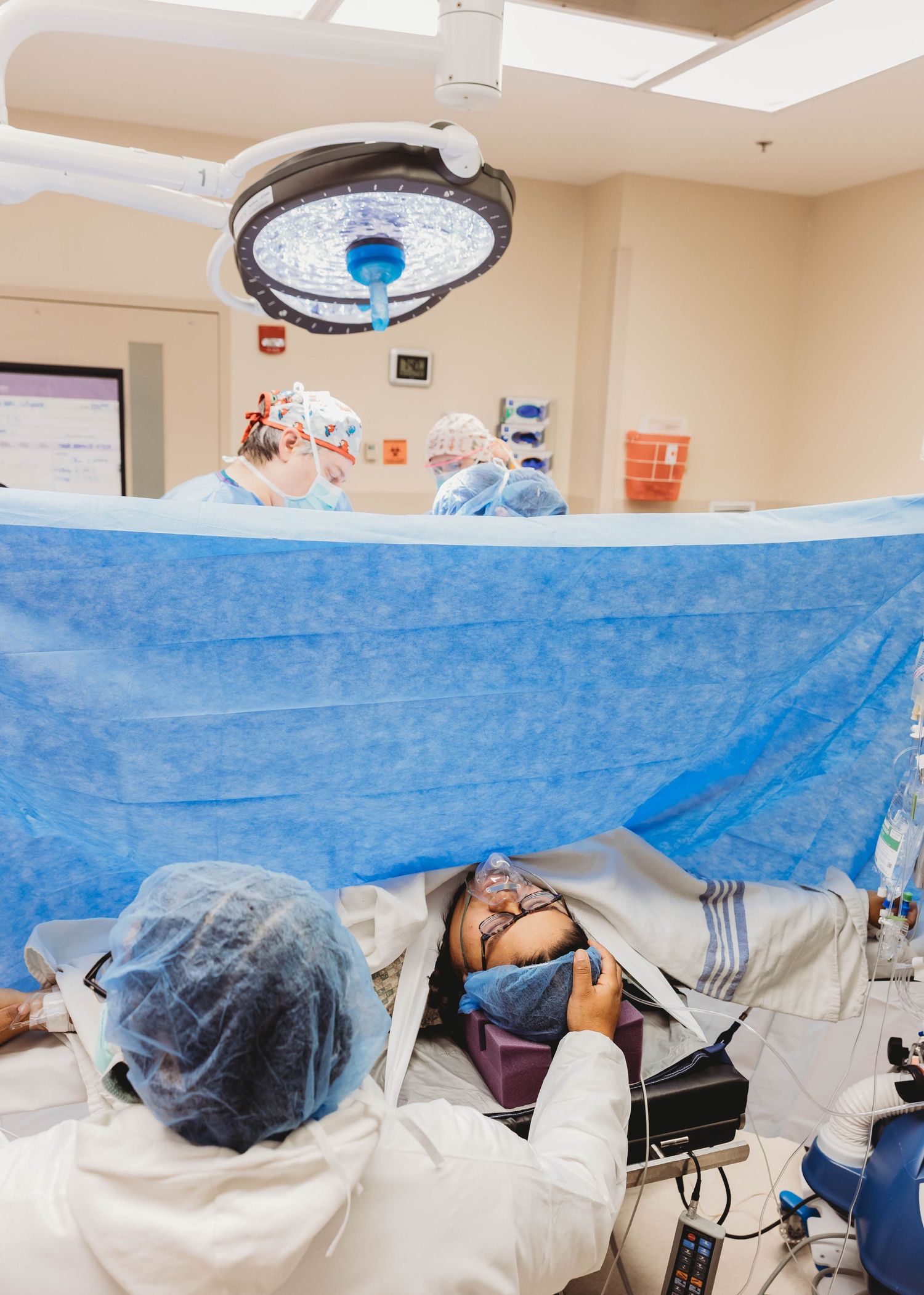 Birth Photographer capturesMedical team performs C-section surgery in operating room with blue drape separating patient from surgical field.
