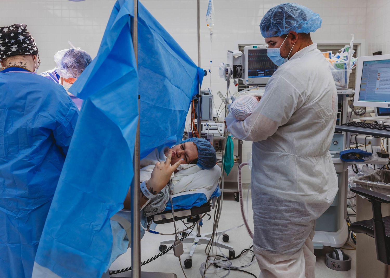 Surgical team in scrubs, masks and caps performs a cesarean delivery as a mother lies awake behind a blue surgical drape.