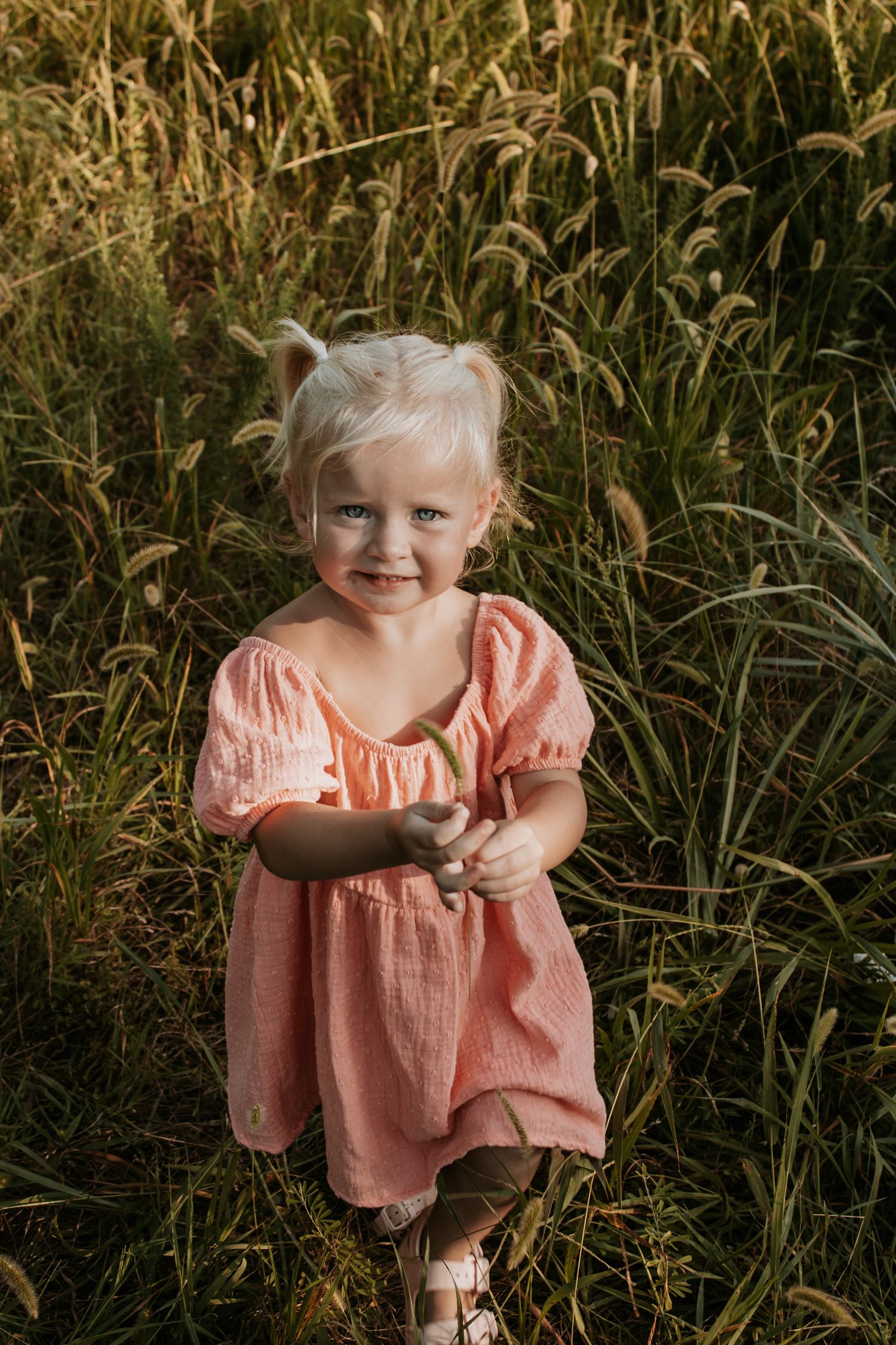 A smiling toddler girl in a pink dress stands in a golden field of tall grass during sunset with Keely King Photography in Claremore, OK