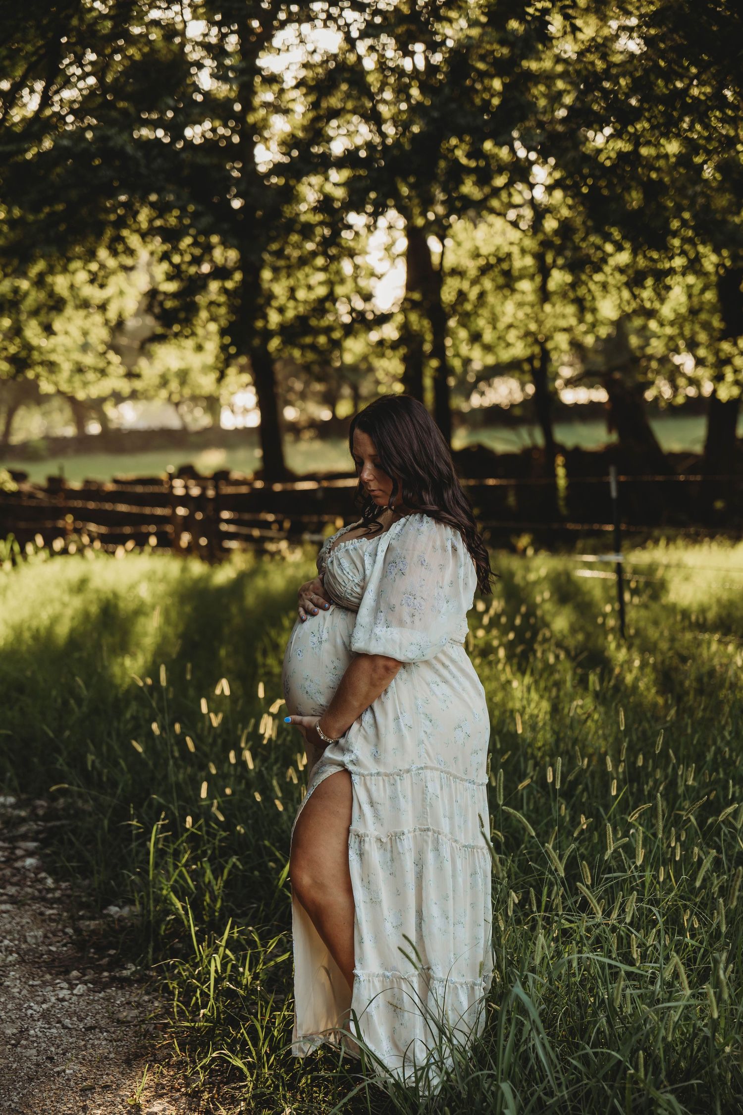 Pregnant woman in white lace dress cradling her baby bump in a sunlit field with a wooden fence behind her.