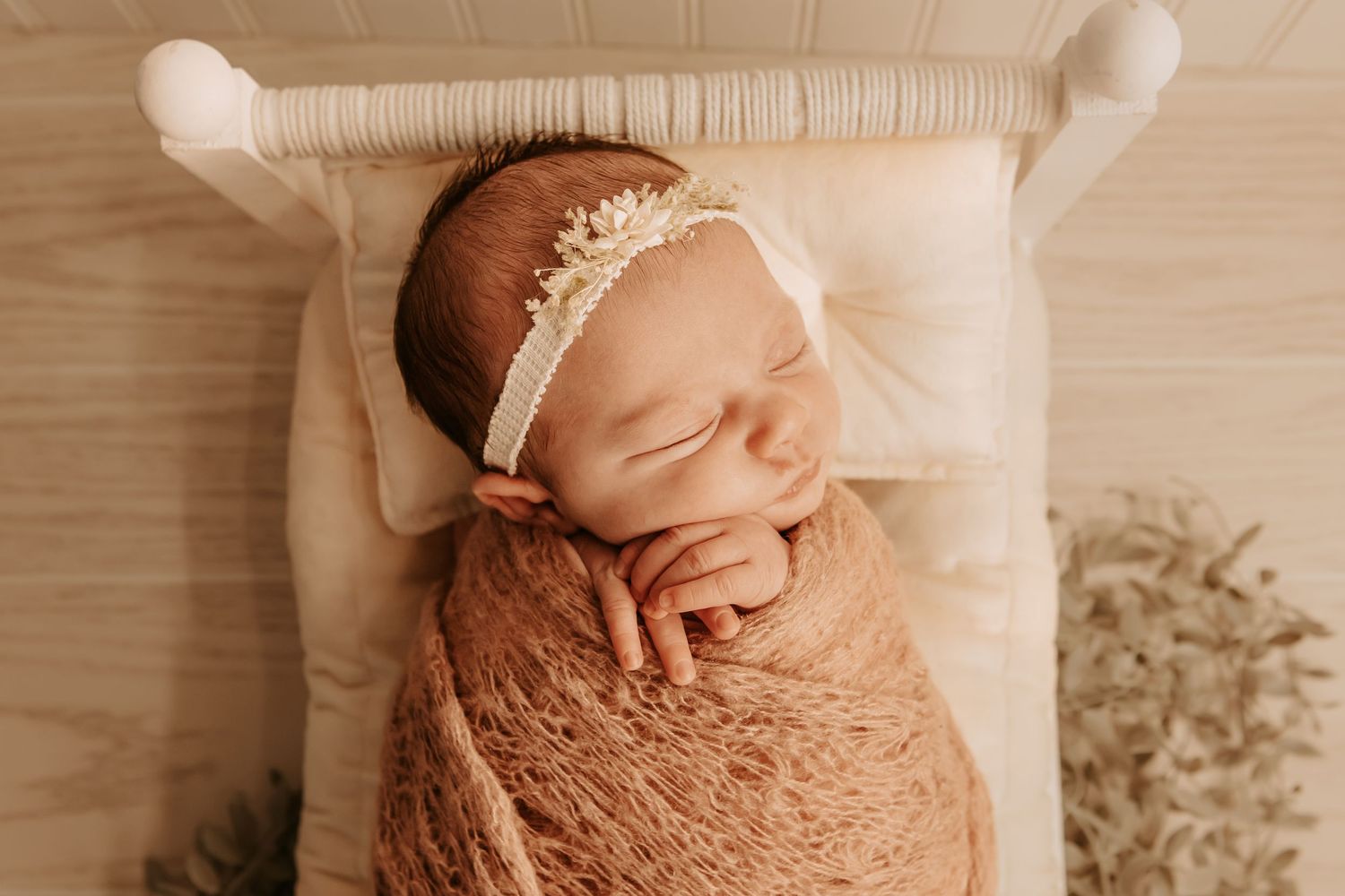 Sleeping newborn baby wrapped in a rust swaddle with a floral headband, resting in a small white wooden bed.