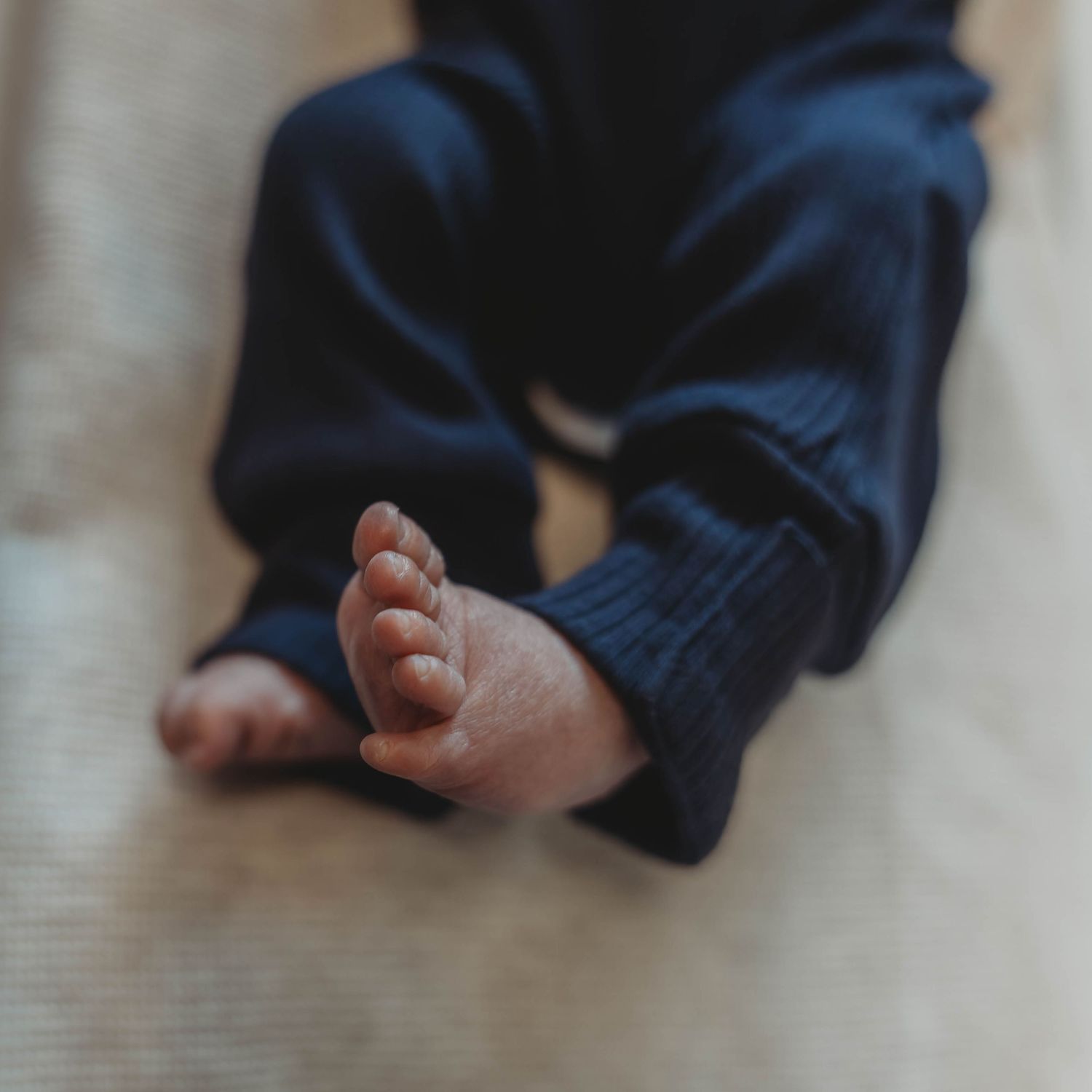 Close-up of tiny baby feet wearing navy blue pants resting on a light wooden floor.