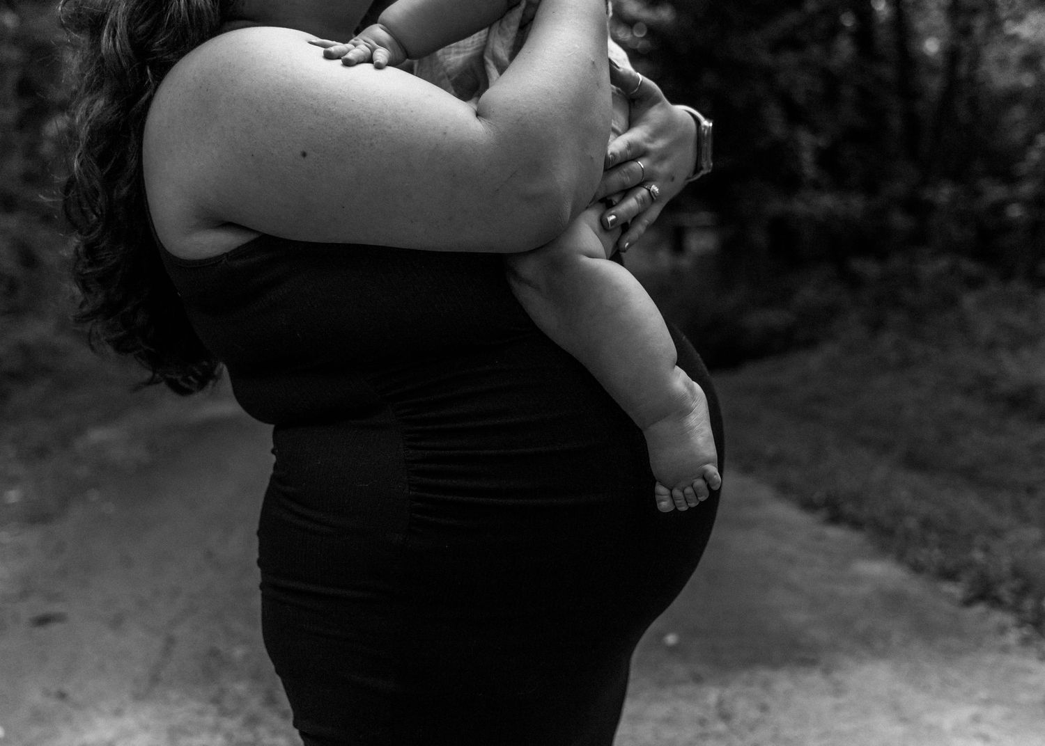 Black and white photo of a pregnant woman in a black dress holding a baby outdoors on a path.