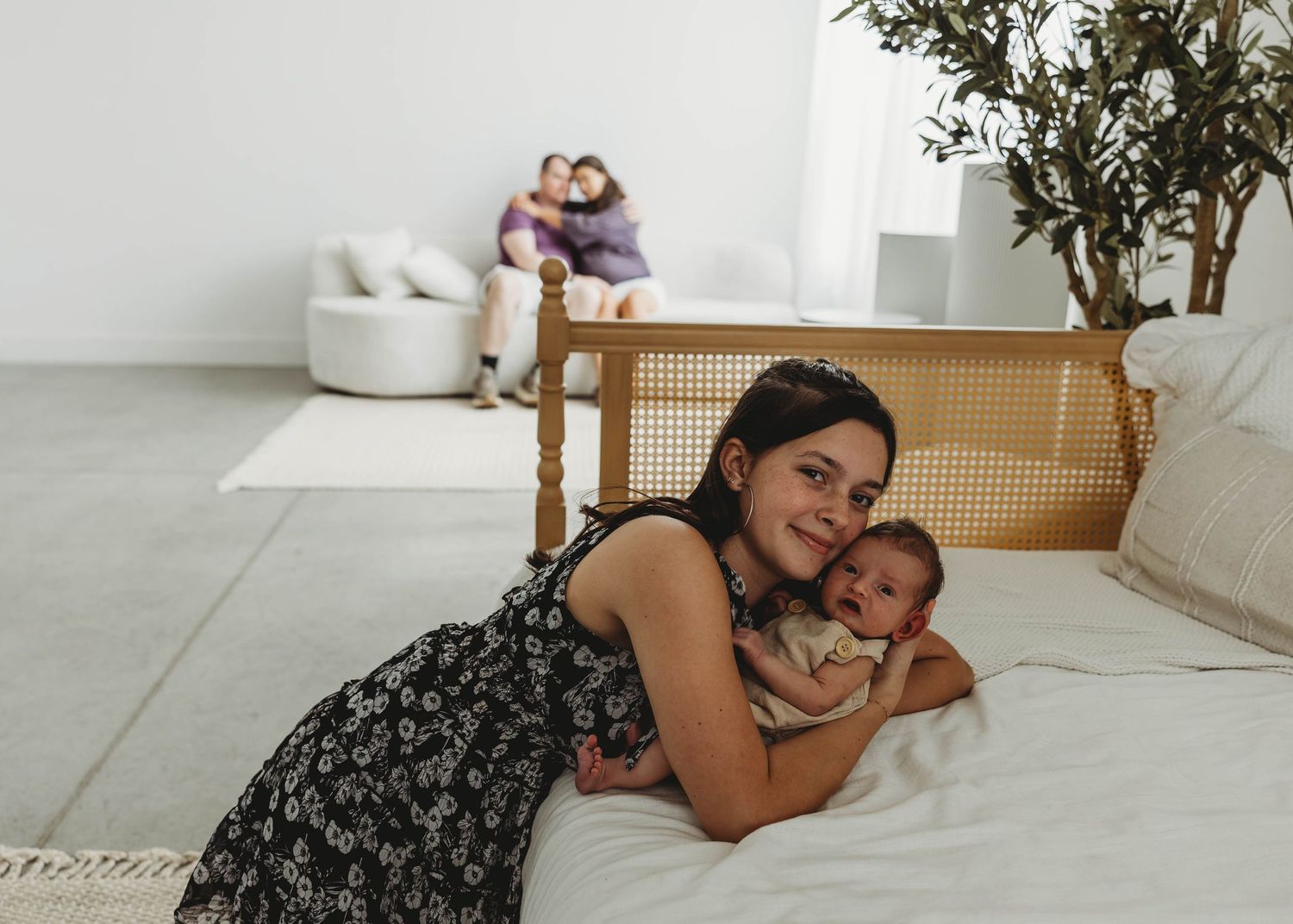 Smiling mother lies with newborn baby on bed while family relaxes in background.