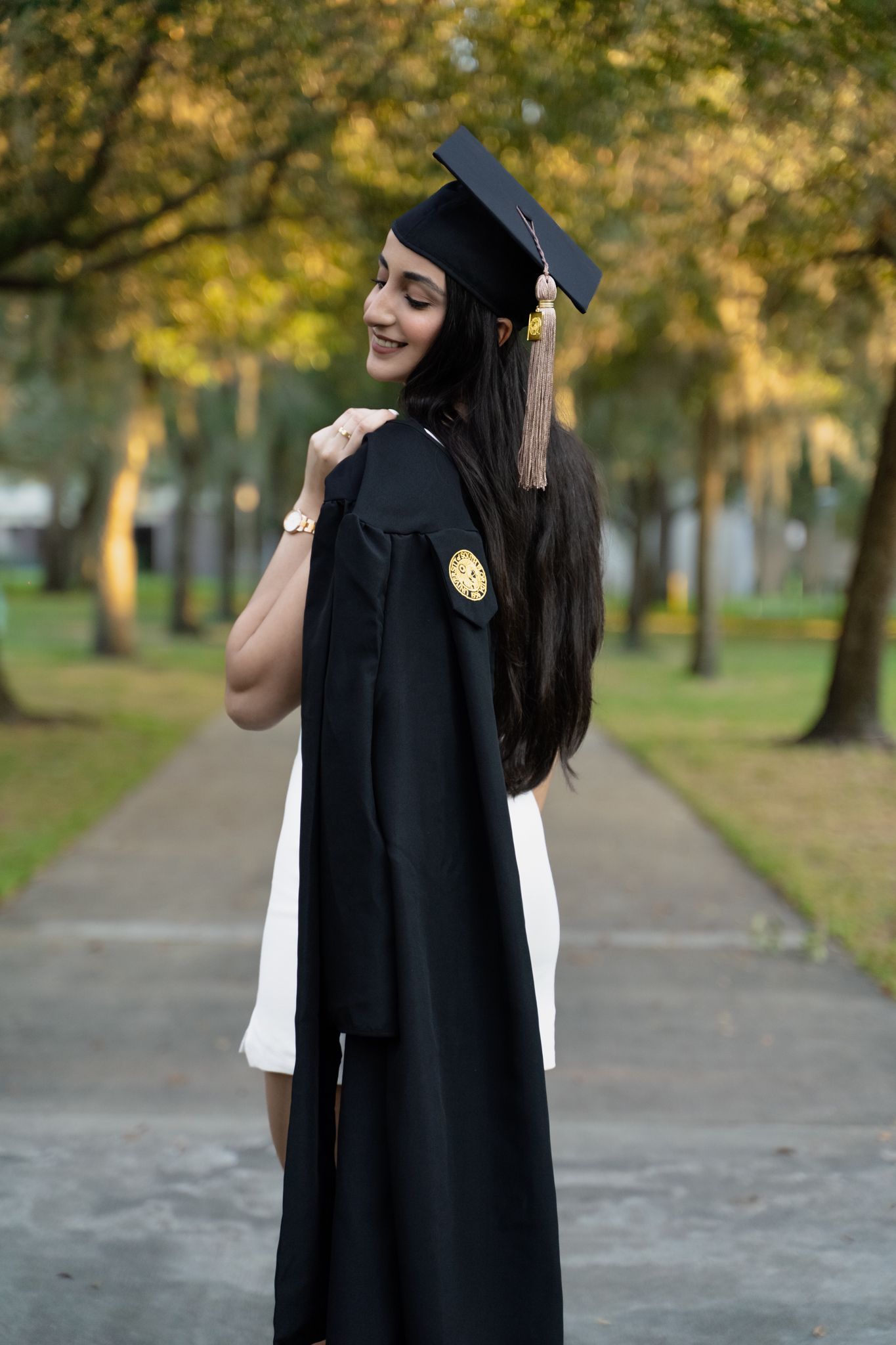 Graduate in black cap and gown smiles over her shoulder on a tree-lined university walkway.