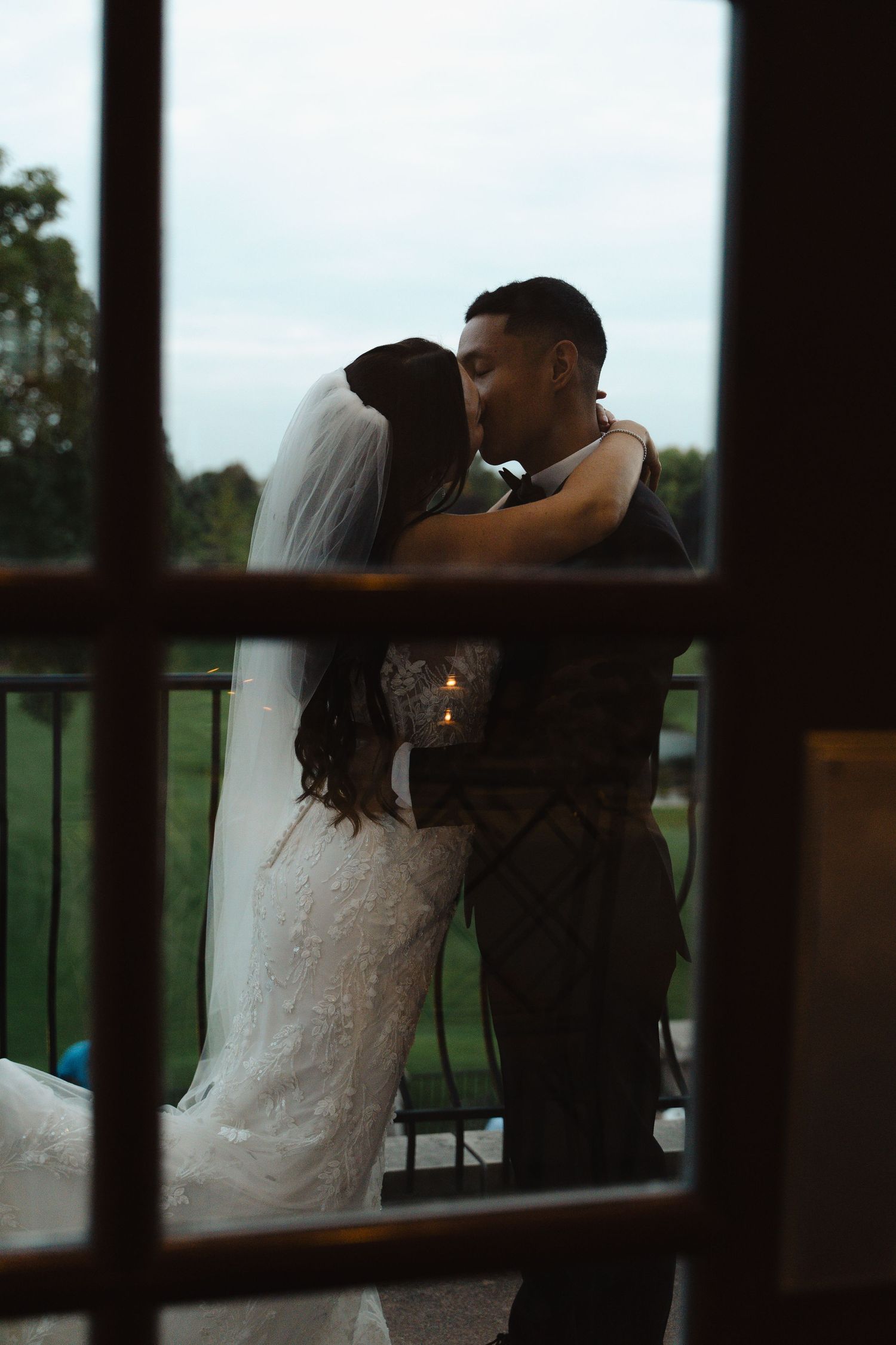 Silhouette of a bride and groom sharing a kiss on a balcony at dusk, viewed through a window pane.