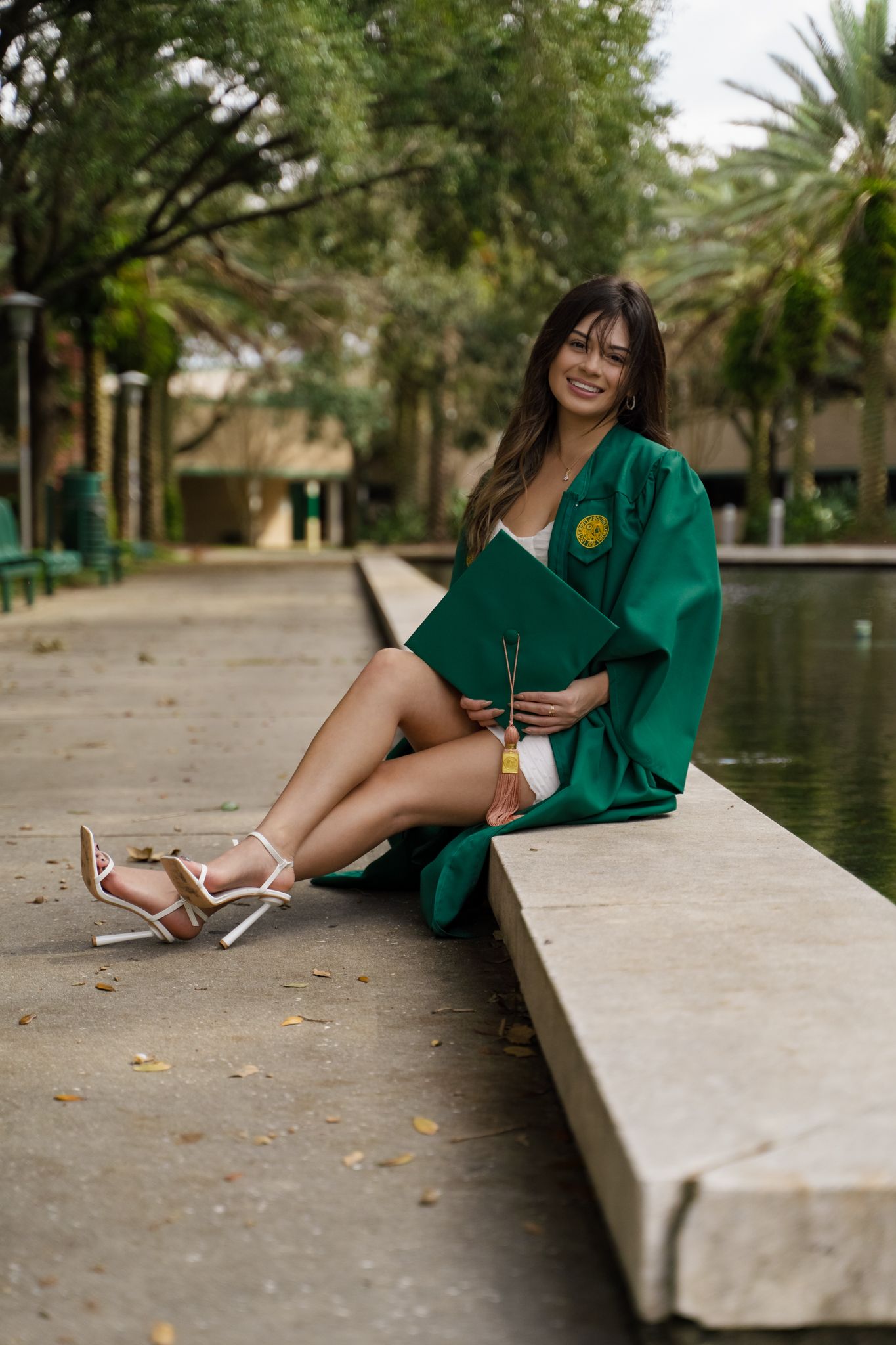 Young woman in green graduation gown sits on stone ledge holding diploma by campus pond.