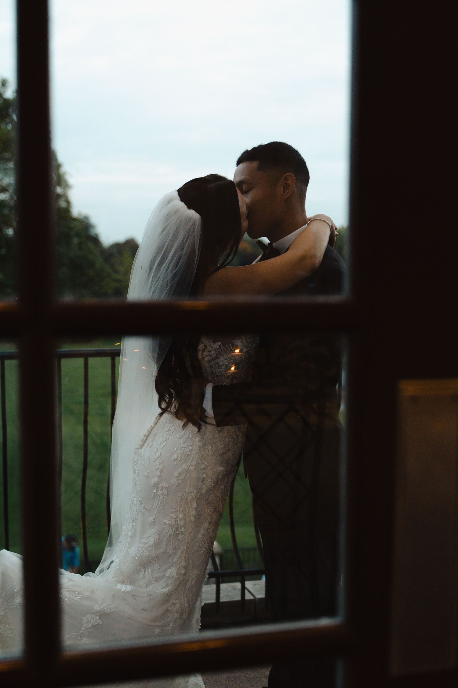 Silhouette of bride and groom kissing on a balcony at twilight viewed through a window.