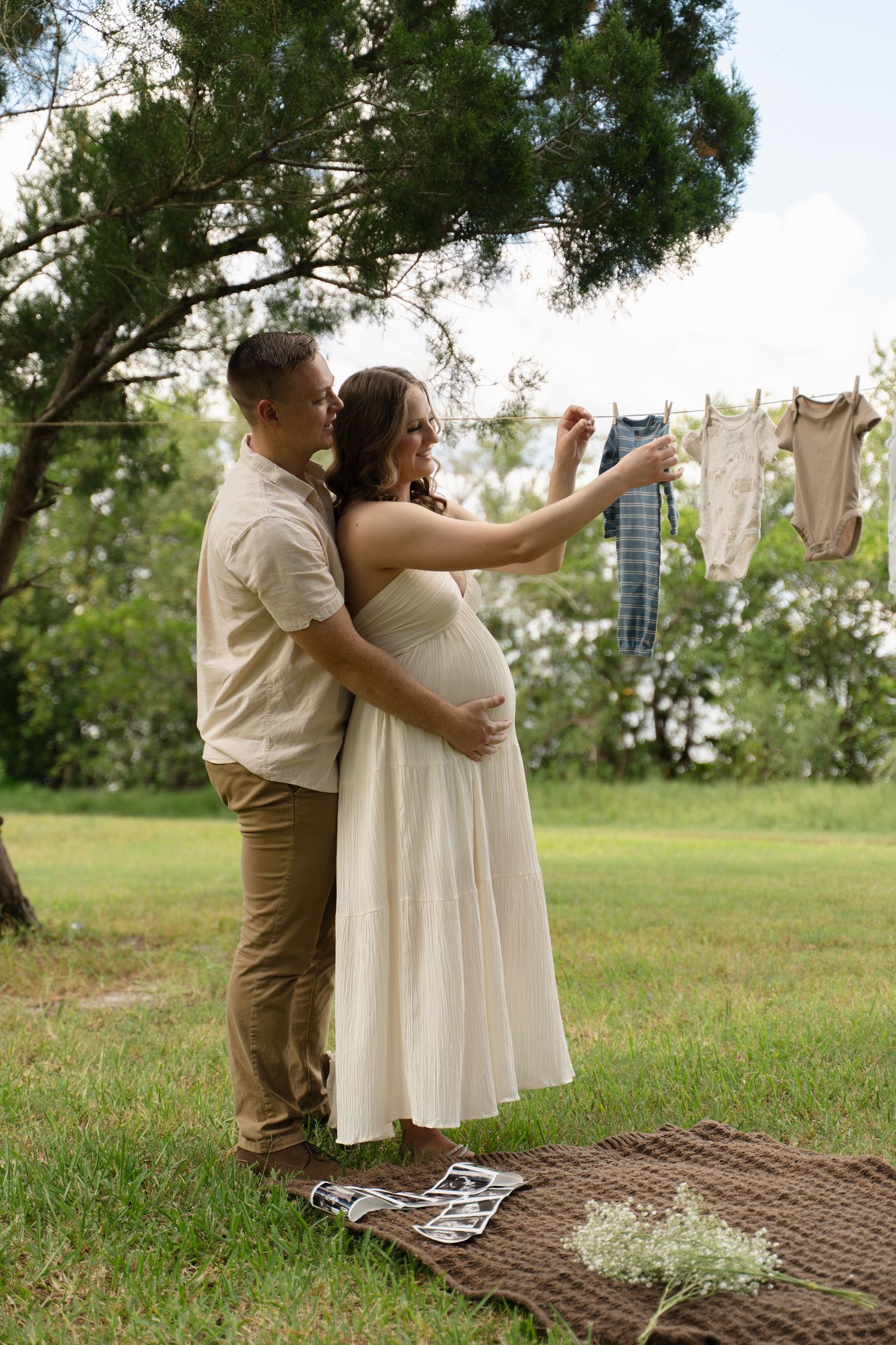 Pregnant couple embracing outdoors near clothesline with baby clothes hanging, woman in white dress pointing at tiny outfits.