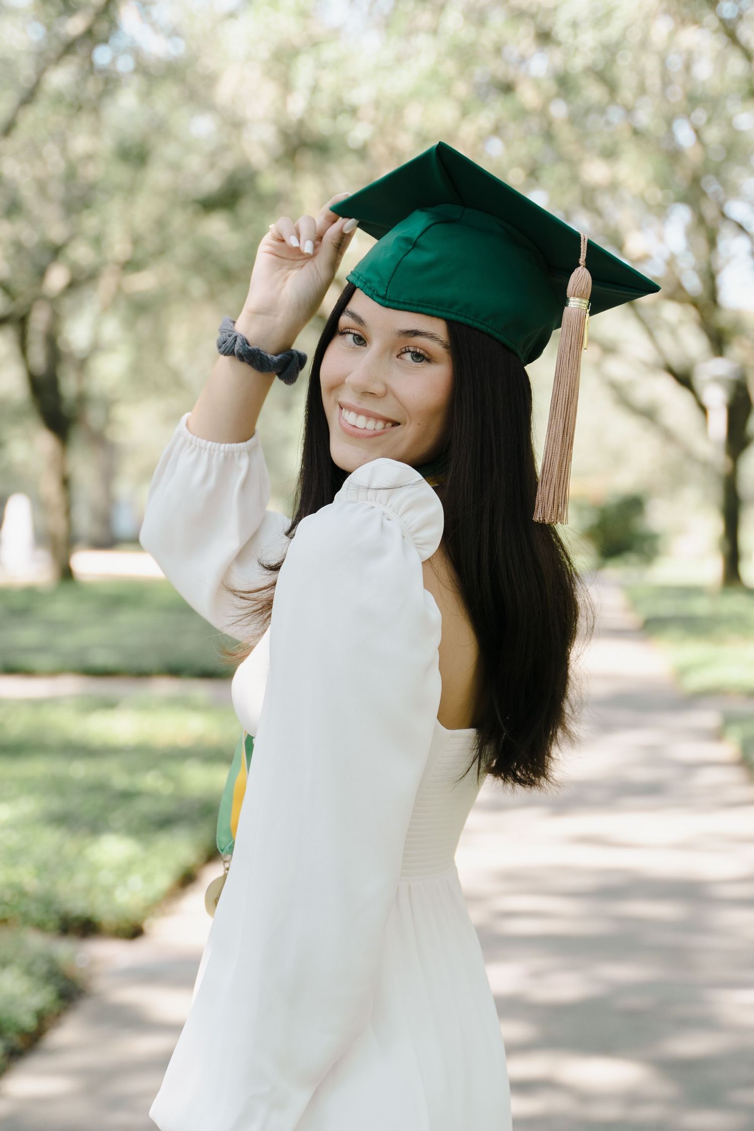Smiling female graduate in white dress adjusting green graduation cap while posing on sunny campus walkway.