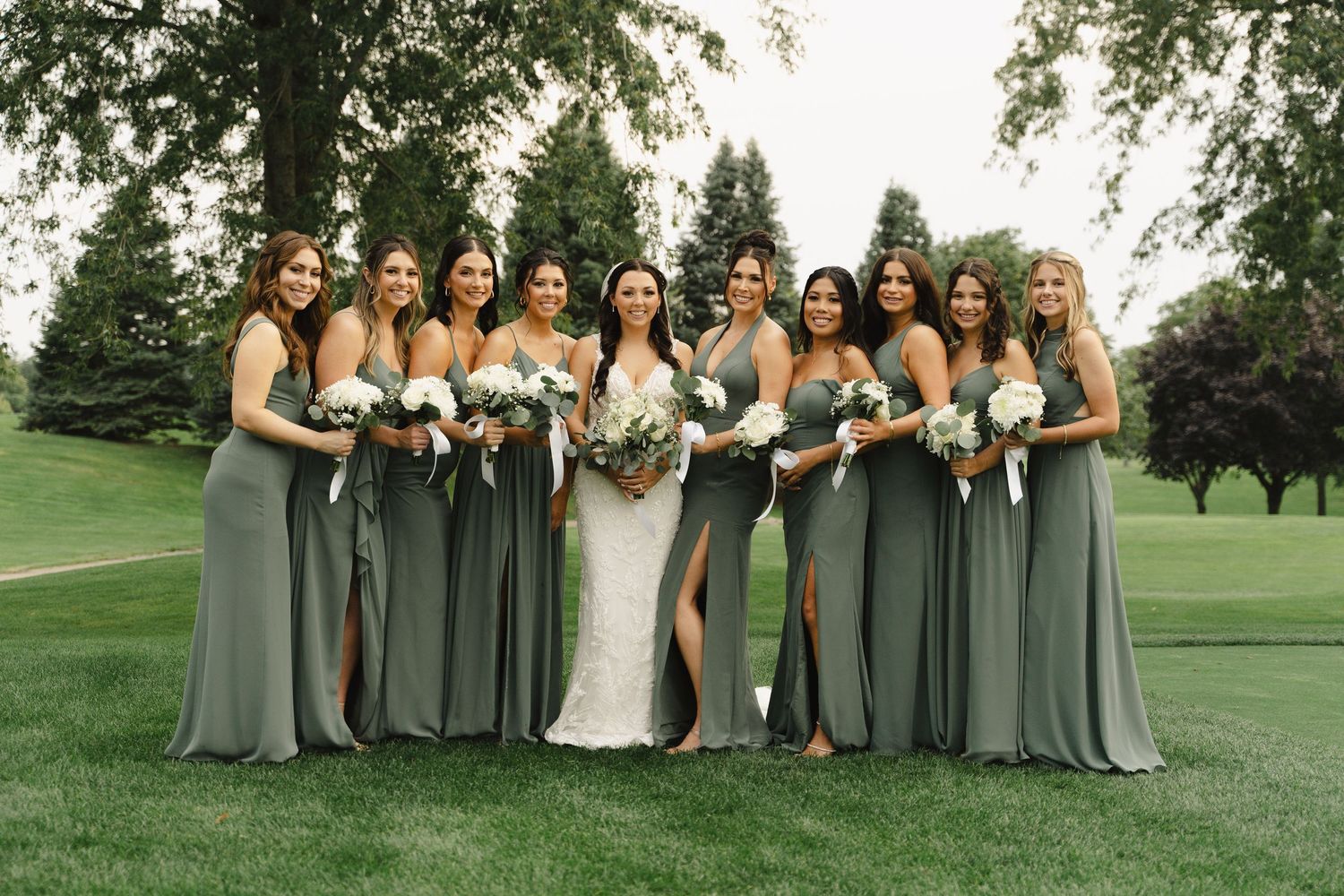 Bride with nine bridesmaids in sage green gowns holding white bouquets on golf course.