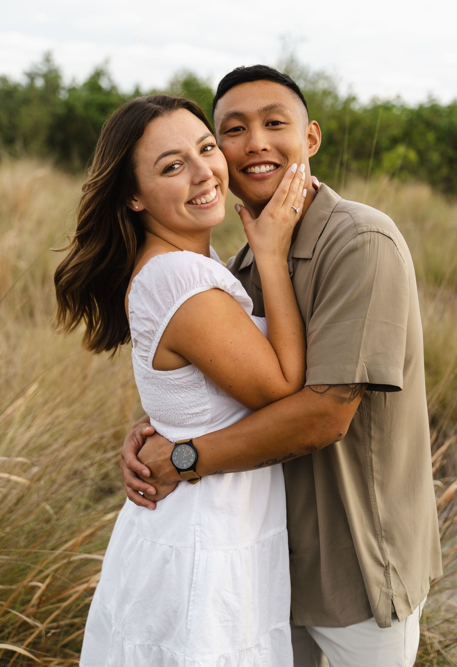A smiling interracial couple embraces in a grassy field, woman in white dress showing engagement ring.
