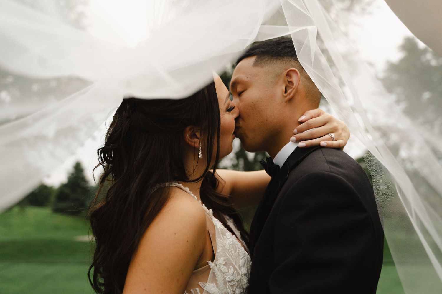 Bride and groom sharing a kiss under a flowing veil on a cloudy wedding day.