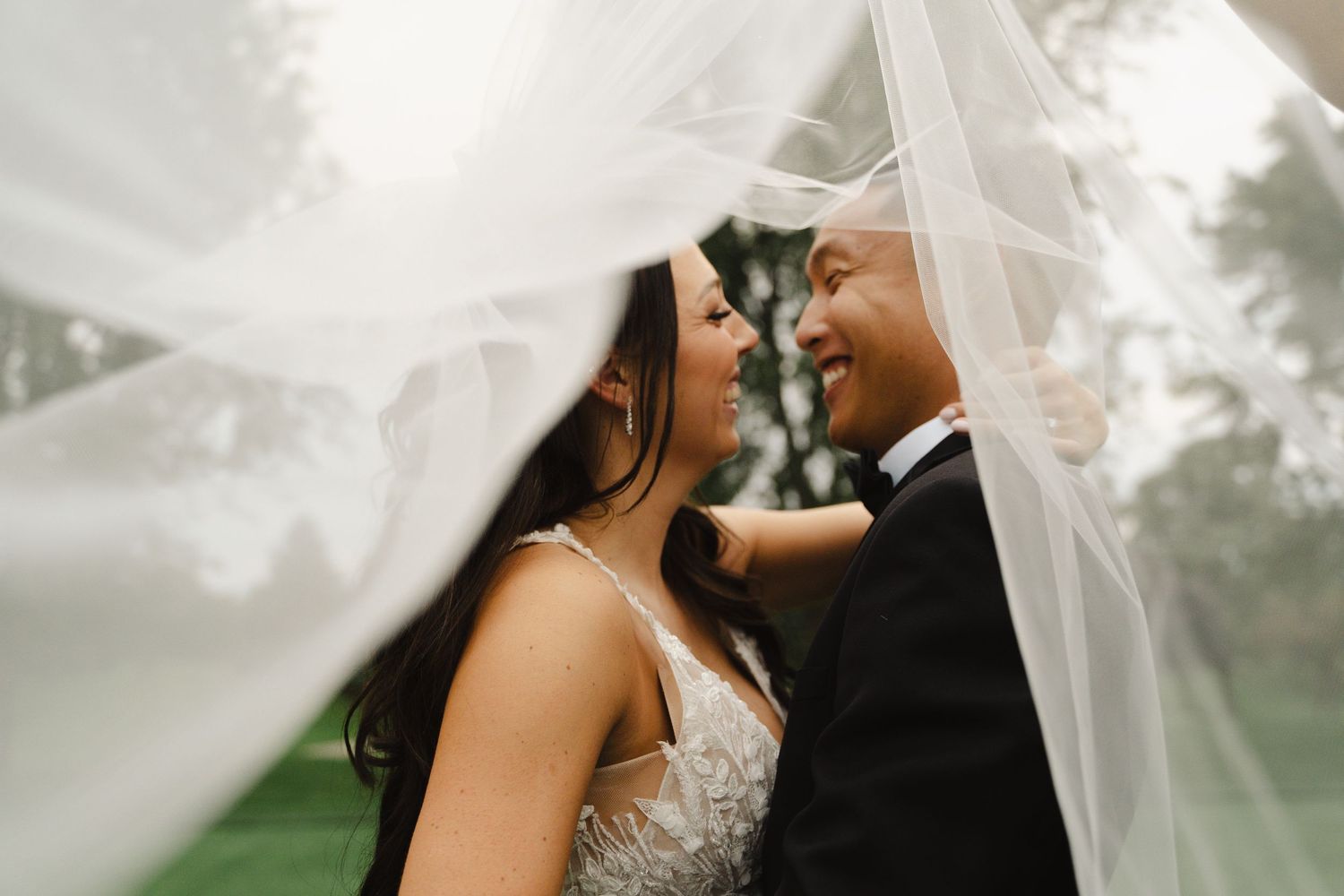 Romantic bride and groom sharing intimate moment under flowing white wedding veil outdoors.