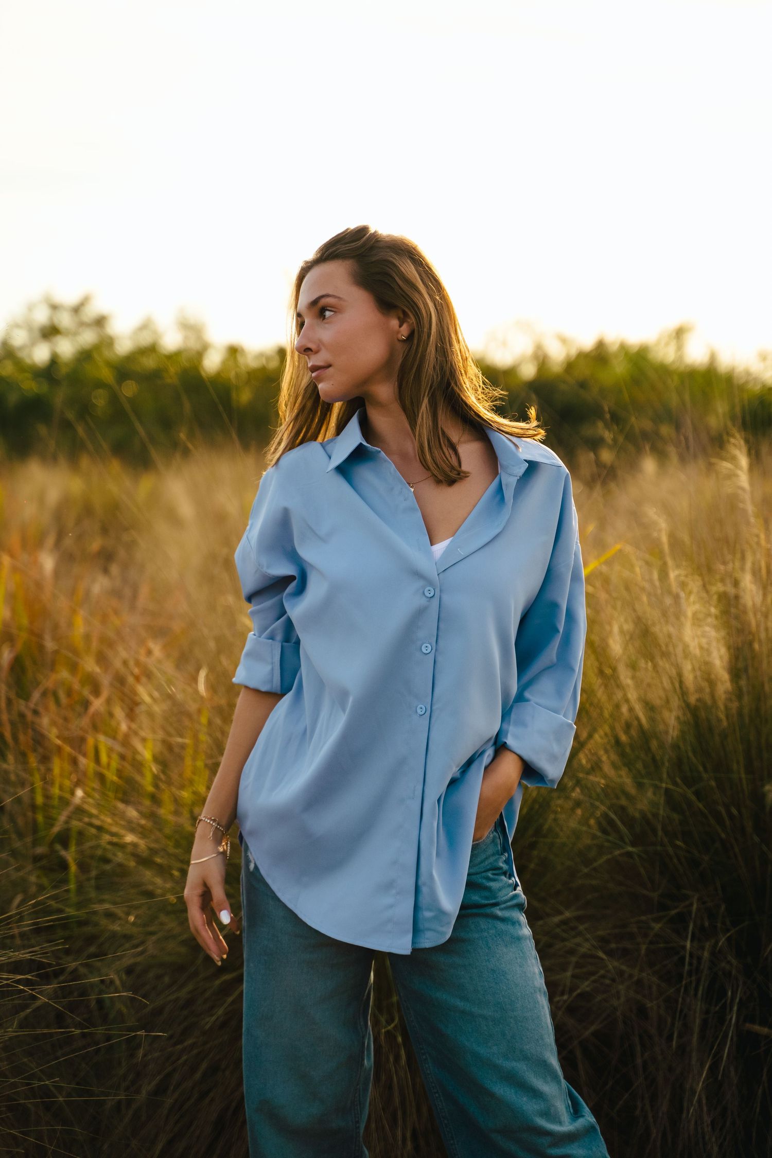 Blonde woman in light blue button-down shirt standing in tall golden grass field at sunset.