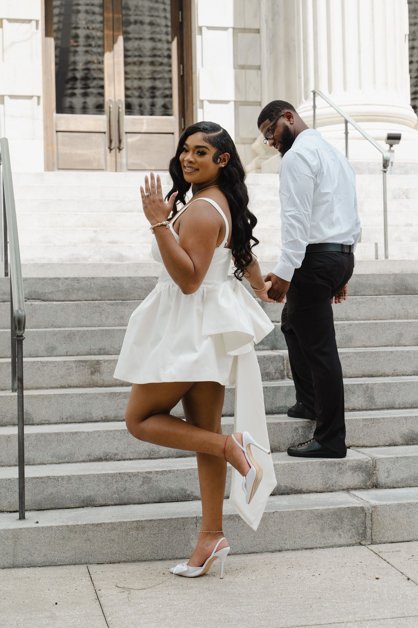 A Black woman in a white peplum dress poses on steps showing her engagement ring while man stands behind.
